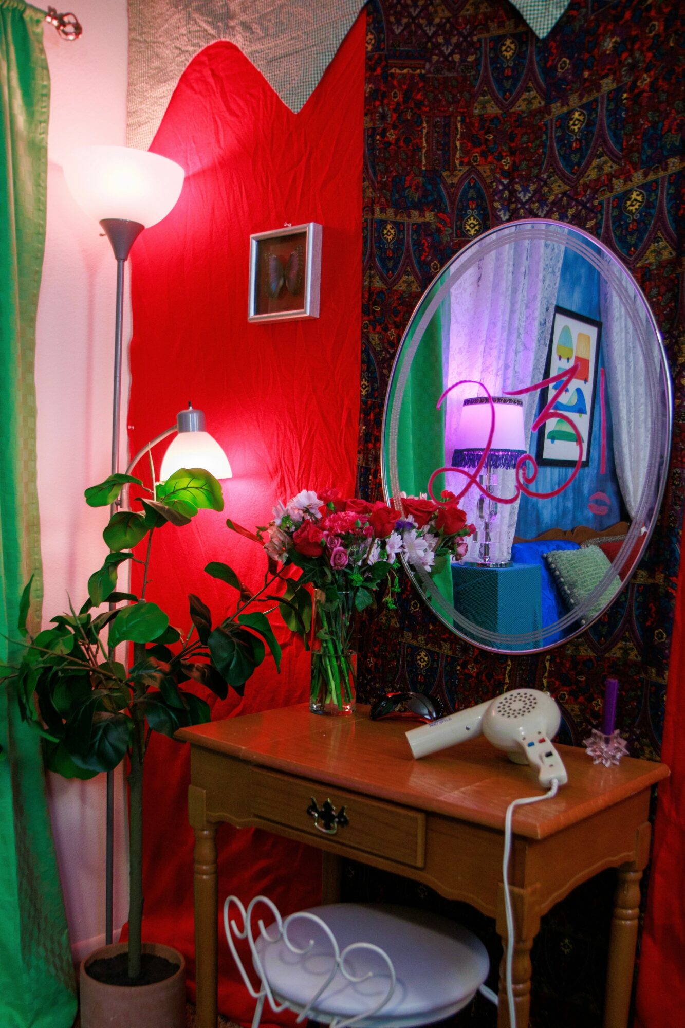 Room with red and patterned walls, mirror, plants, flowers, and a wooden table with a hairdryer and phone.