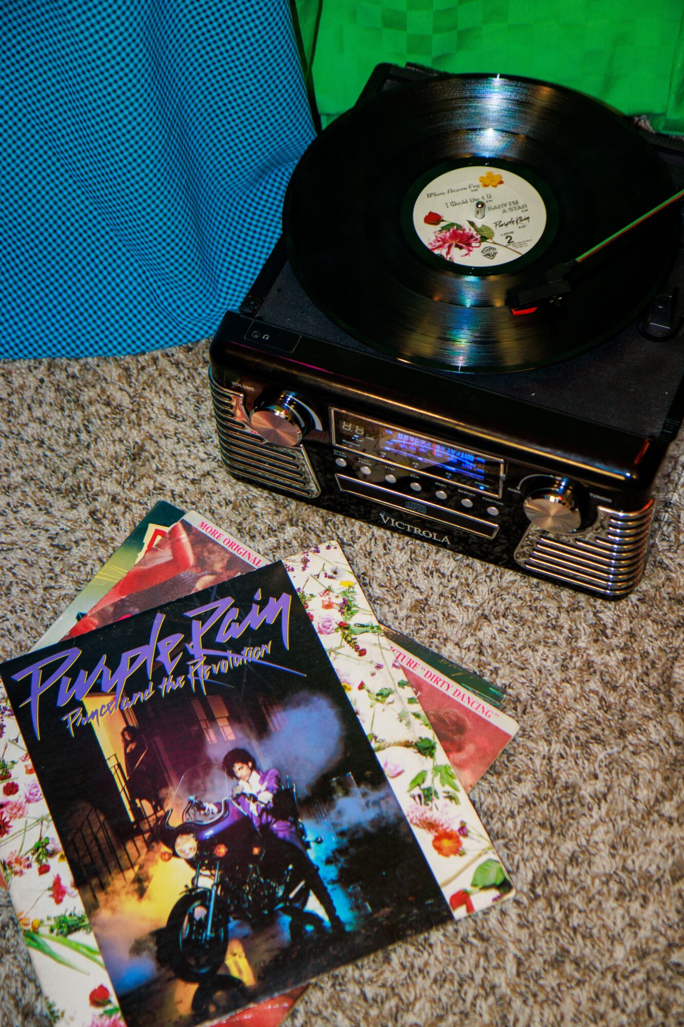 Turntable playing vinyl record with colorful album covers on carpeted floor, green and blue background.
