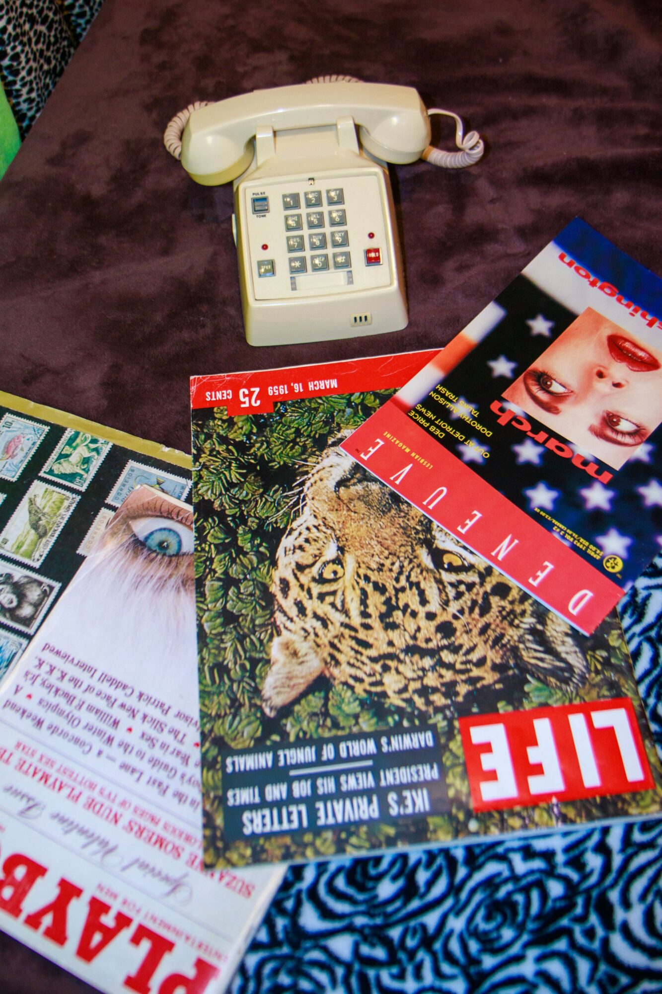 A vintage white rotary telephone, magazines with images of a leopard and a woman, and a blue patterned fabric surface.