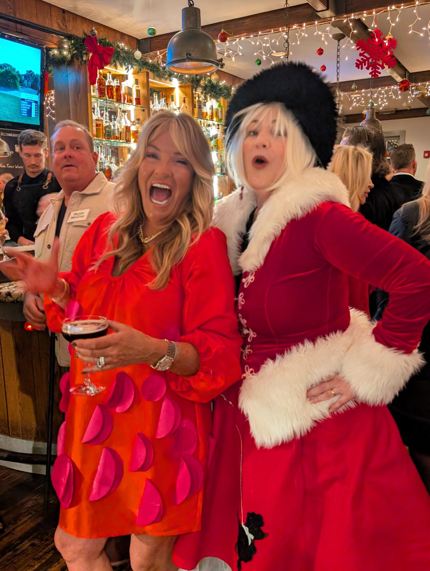 Two women in festive red outfits smiling and posing in a decorated bar with holiday lights and decorations.