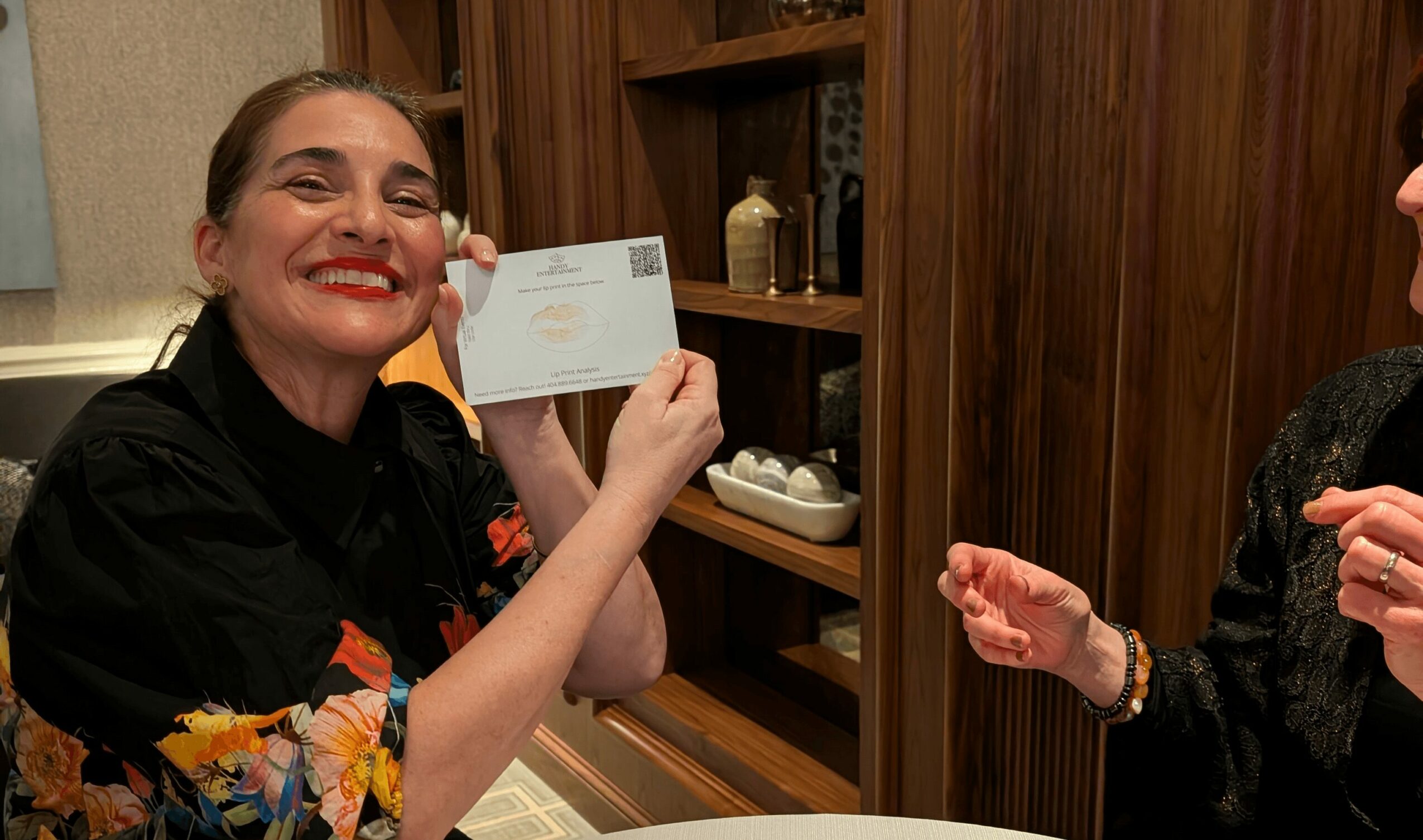 Woman smiling and holding an envelope, with another person partially visible, in a room with wooden shelves.