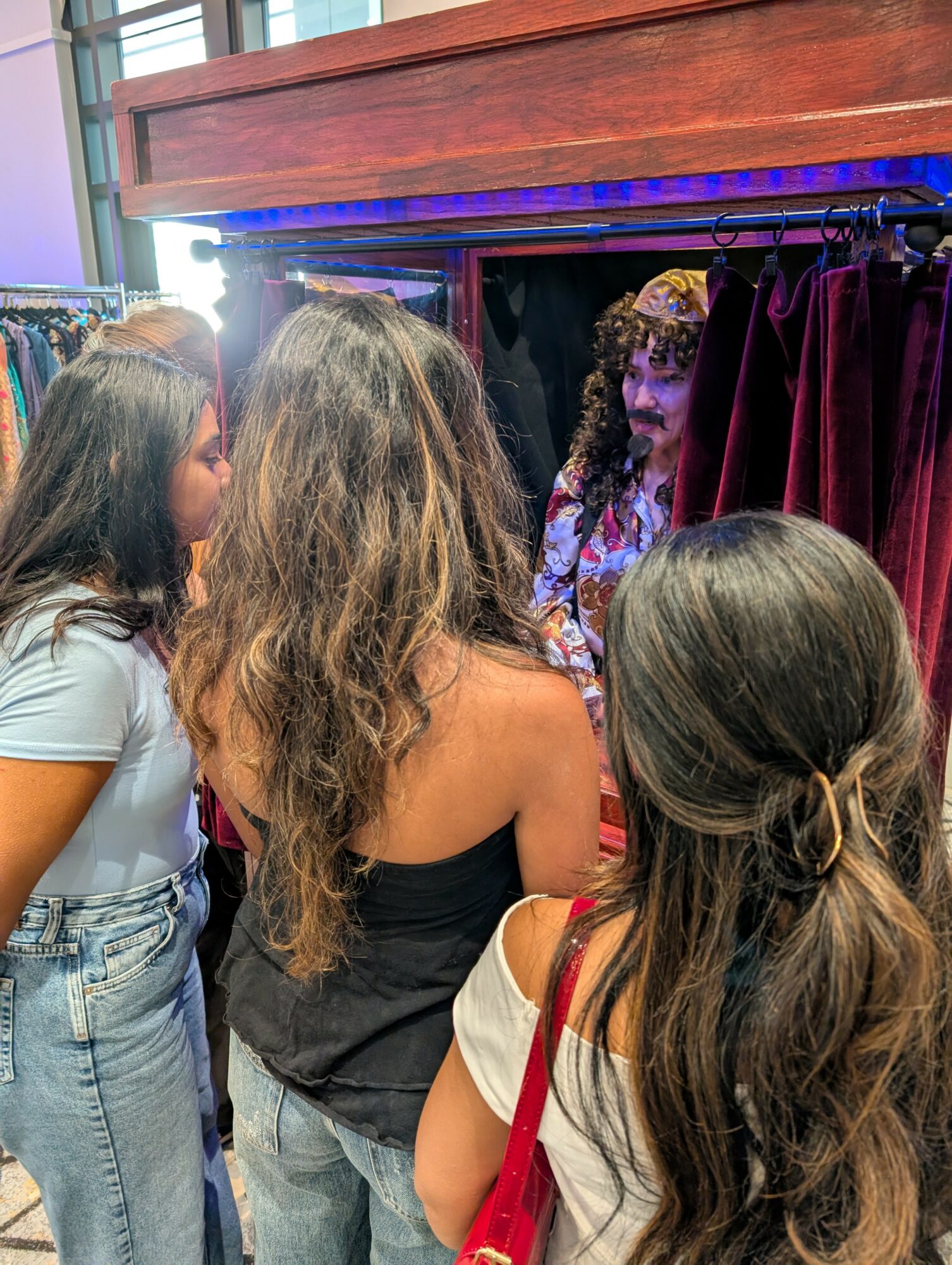 Four women are gathered around a small stage with a curtain, engaging with a performer dressed in costume with curly hair.