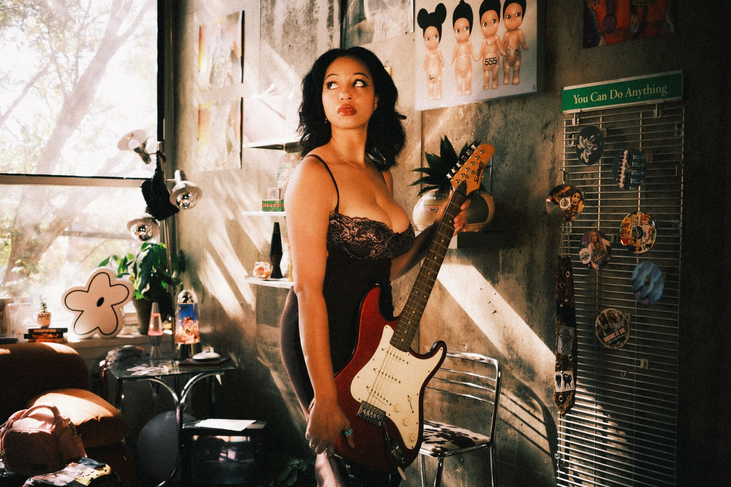 Woman with dark hair holding an electric guitar in a room with posters and sunlight streaming in.