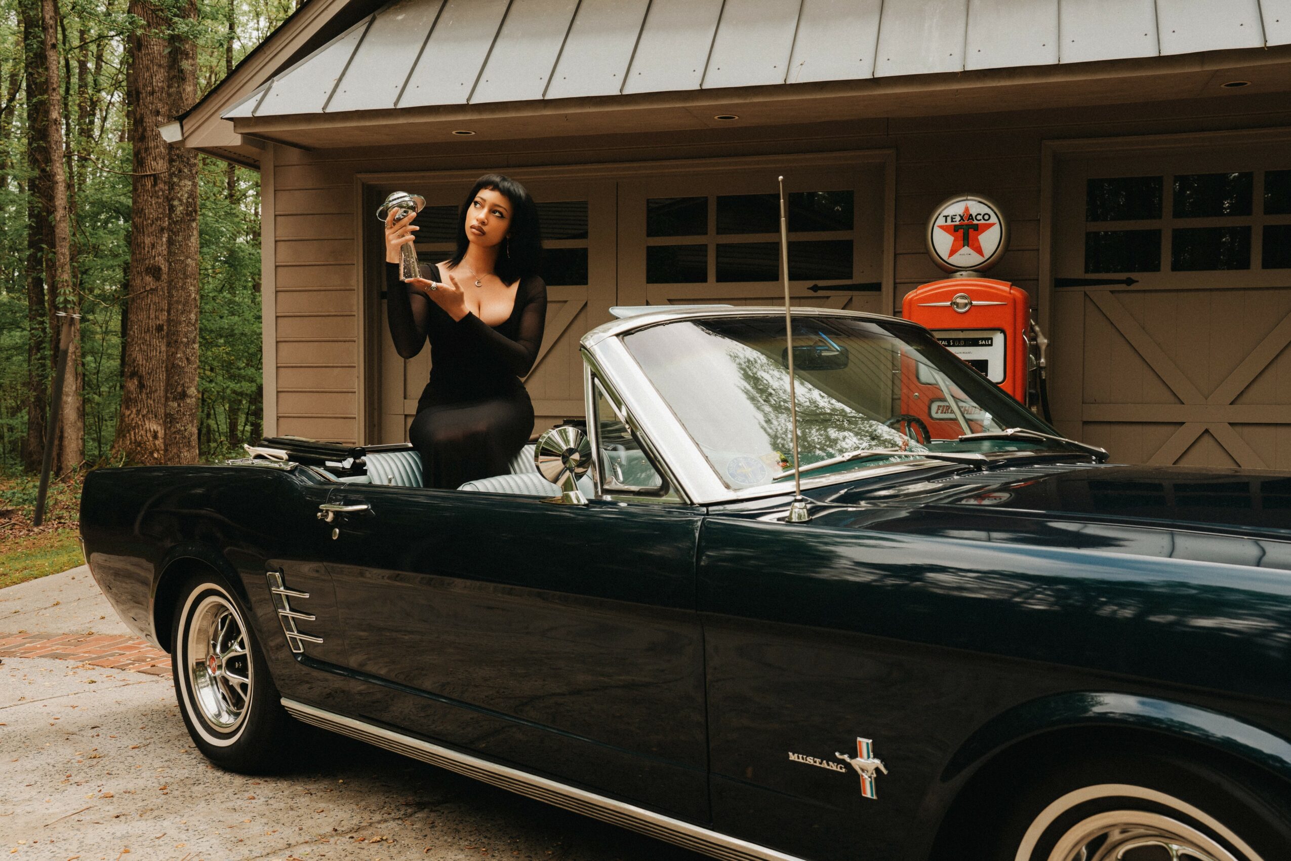 Woman in black dress standing next to a black vintage convertible car, holding a drink, in front of a garage with trees nearby.