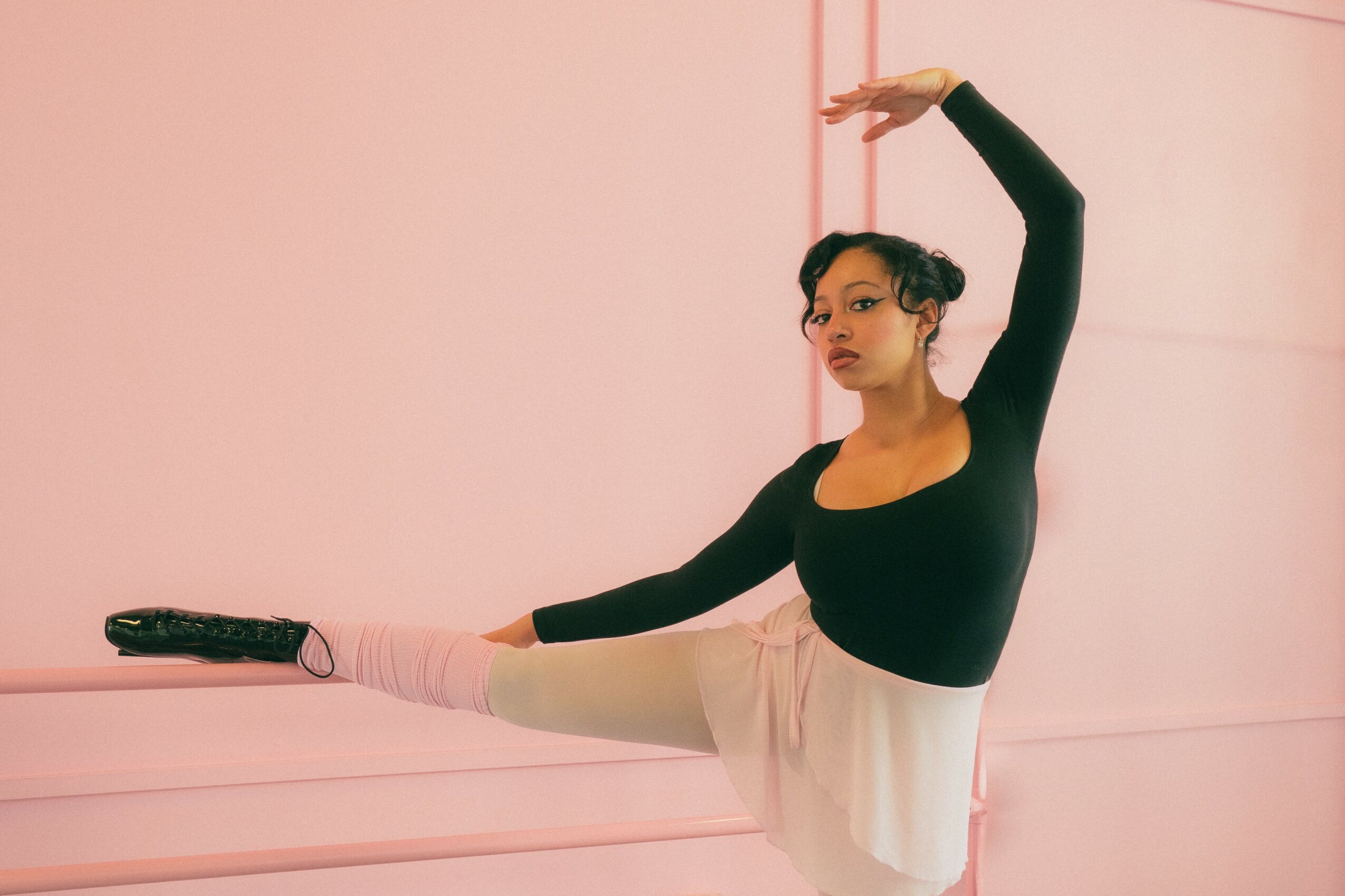Ballet dancer in black top and beige skirt stretches leg on pink wall, raising arm overhead, in dance studio.