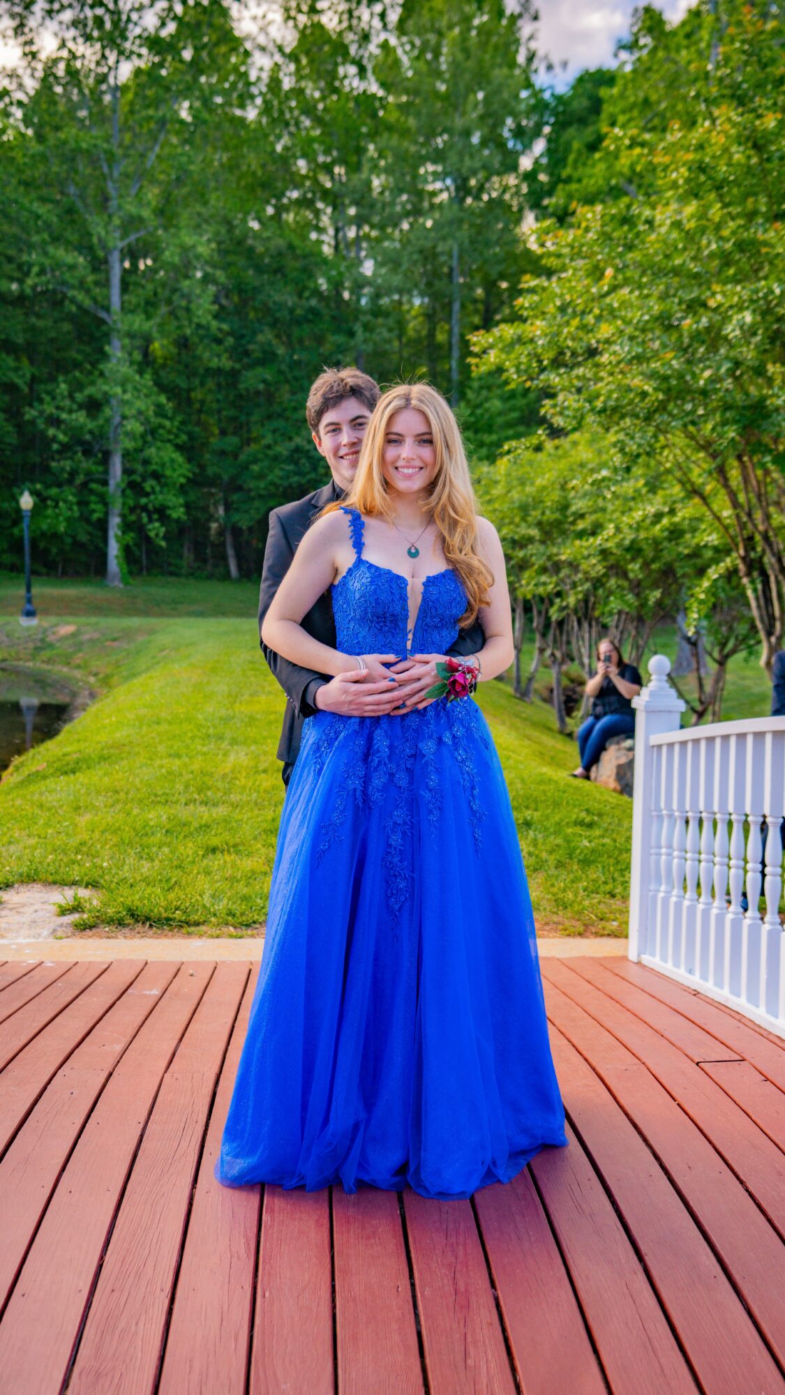A young woman in a blue gown stands on a wooden deck outdoors, with a man behind her and trees in the background.