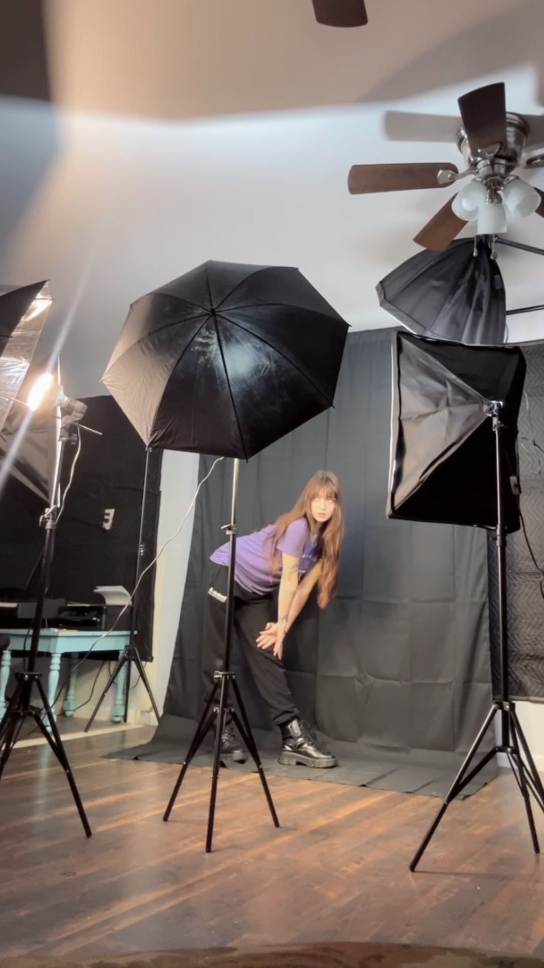 Person with long hair in purple shirt bending forward in a photography studio with lighting equipment and black backdrop.