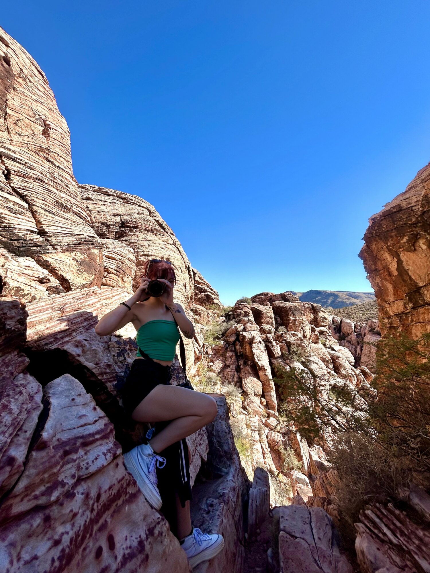 Person sitting on rocks in a canyon with blue sky above, wearing a green top, black shorts, and white sneakers.