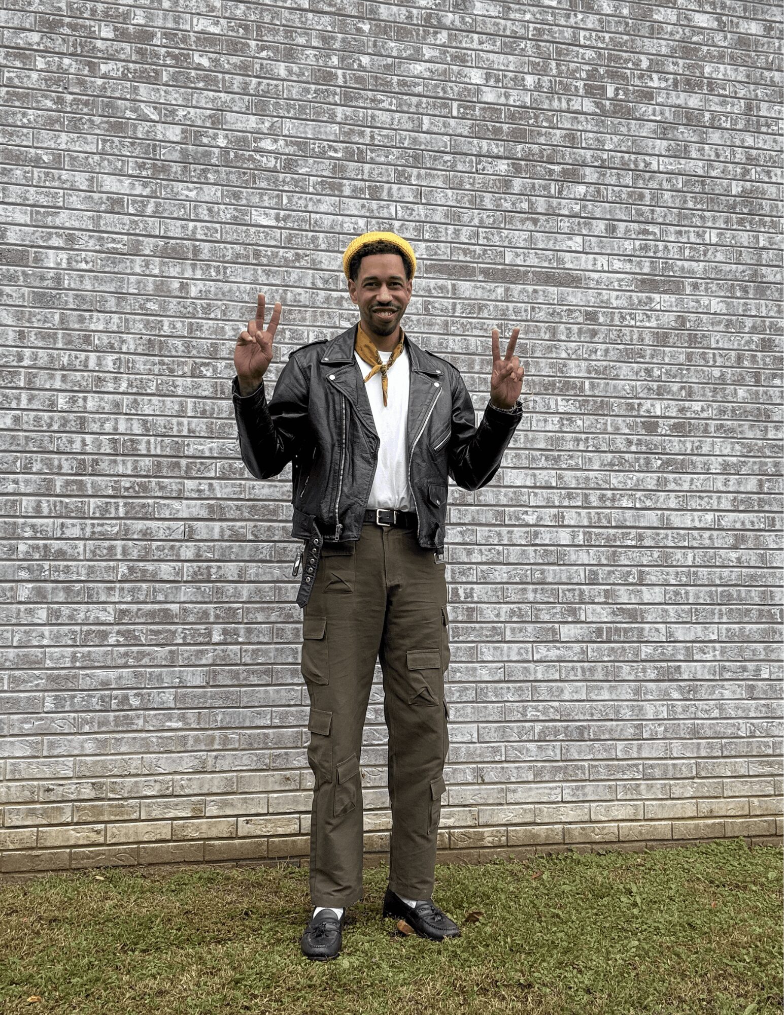 Man standing outdoors in front of a brick wall, making peace signs with both hands, smiling, wearing a leather jacket and yellow hat.