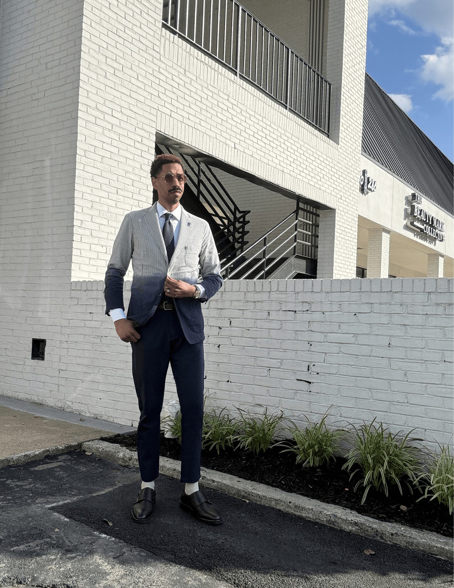 Man in suit holding a folder standing outside near a white brick building with stairs and a balcony.