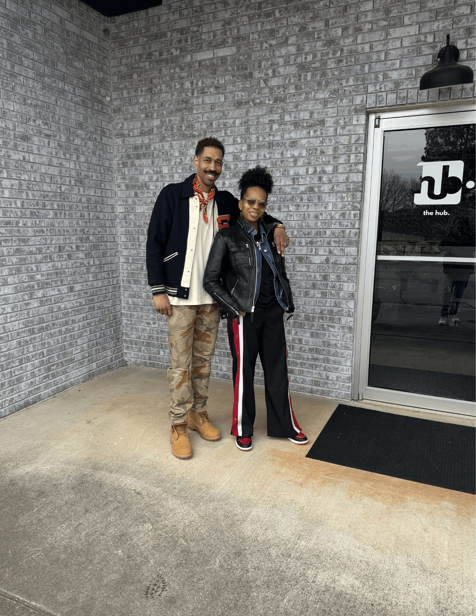 Two people standing outside near a brick wall and glass door, smiling, with one person having arm around the other.
