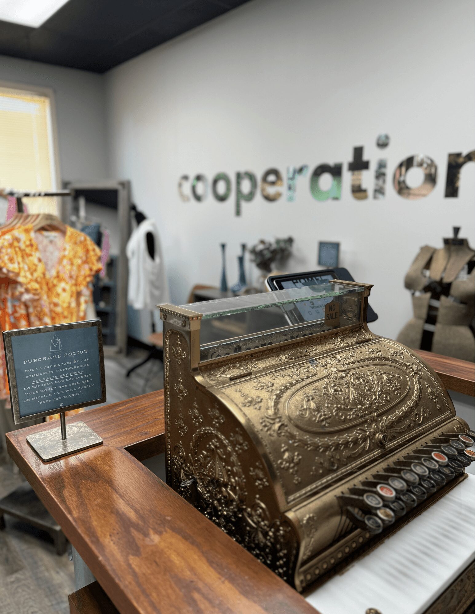 Cash register on a wooden counter in a store with clothing and a sign in the background. The store has a white wall with colorful letters spelling 'cooperation'.