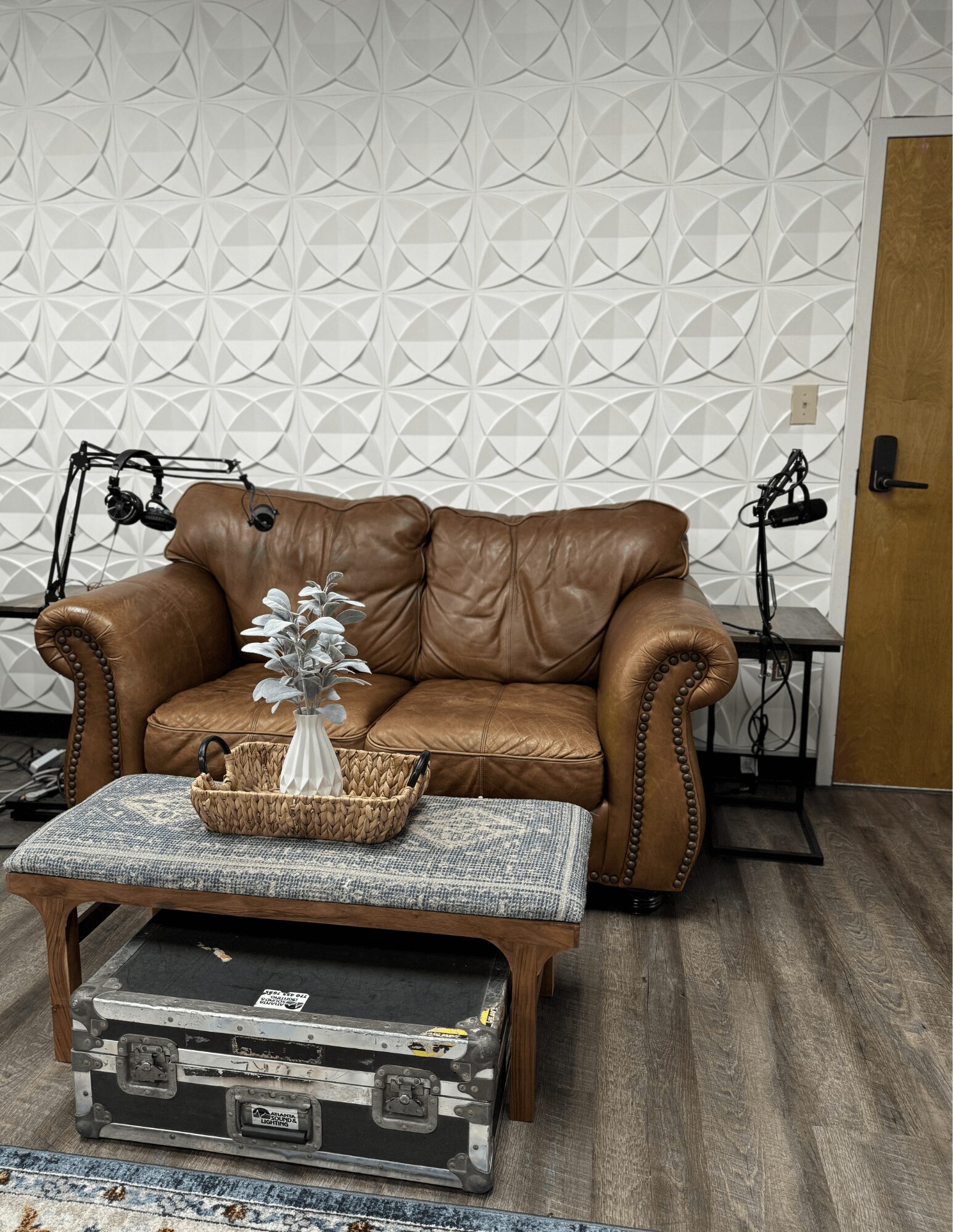 Brown leather sofa with a vase of flowers and a basket on it, against a textured white wall, with a wooden door to the right.