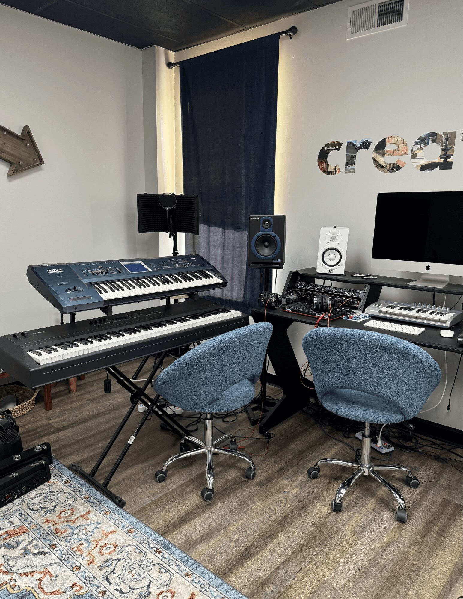 Music studio with keyboards, speakers, computer, and two blue chairs in a room with dark curtains and a decorative rug.