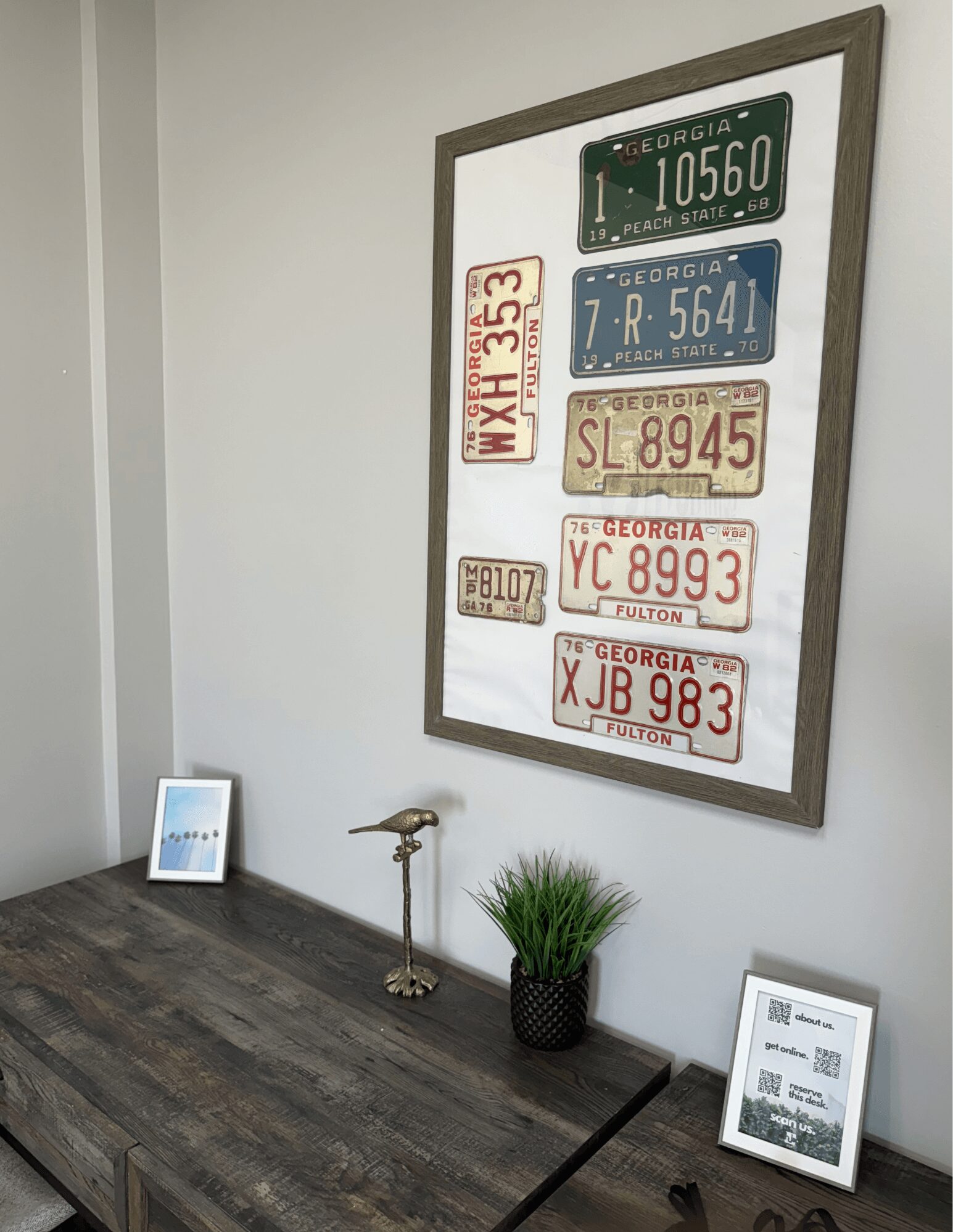 Wall with framed license plates, small framed picture on table, brass lamp, potted plant, and framed document, all on a wooden surface.