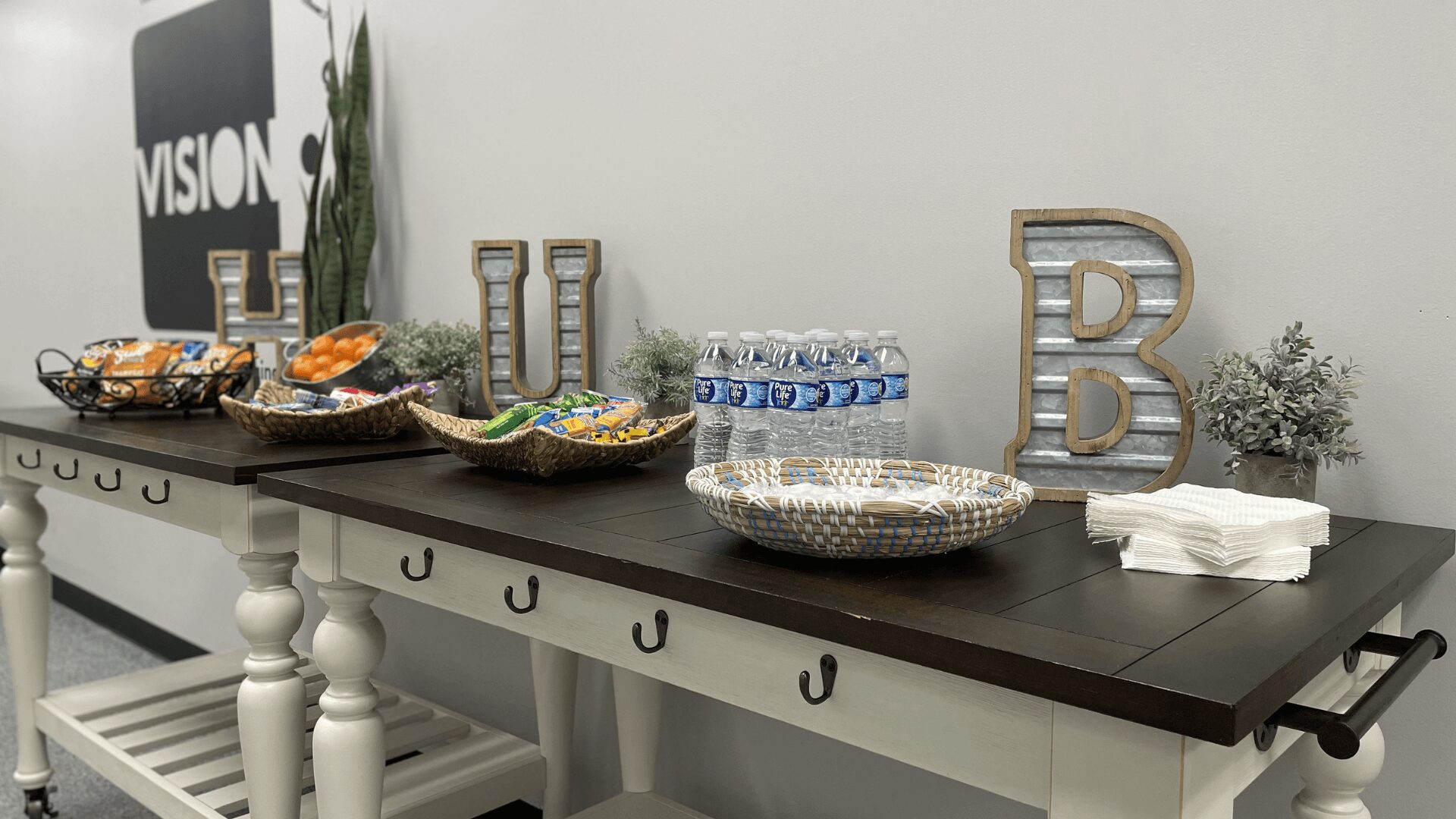 Decorative table with snacks, water bottles, and letter decorations, against a white wall with a plant and framed art.