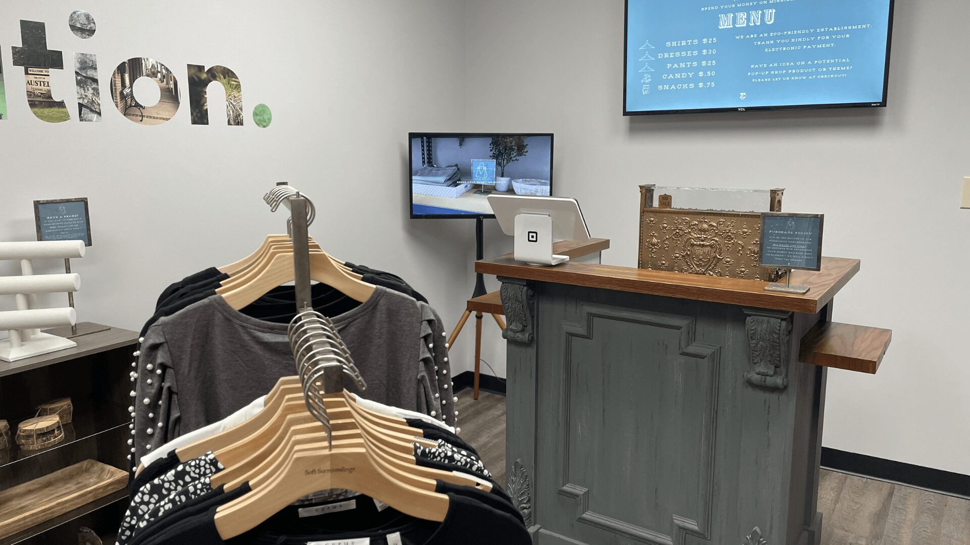 Clothing display with hangers and shirts, checkout counter with computer, and wall screens in a retail store.