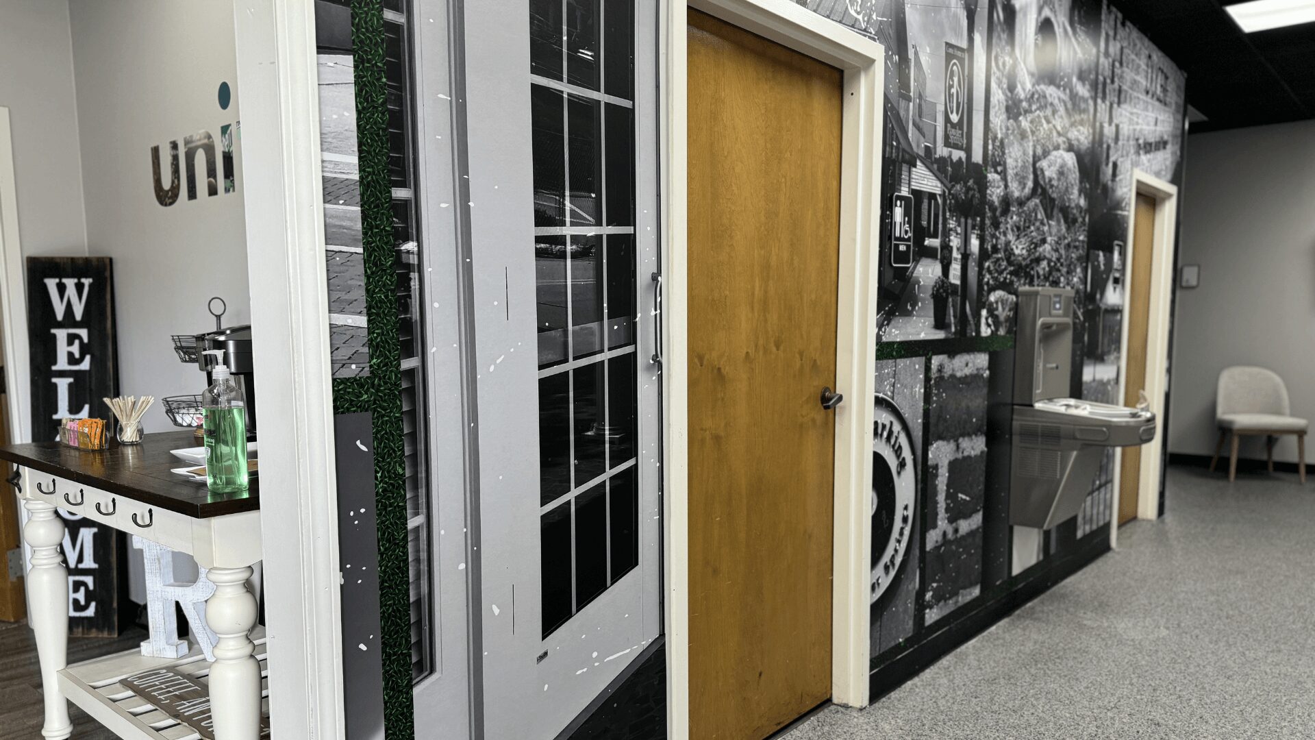 Interior hallway with a wooden door, black and white wall art, and a small sink area with a chair nearby.