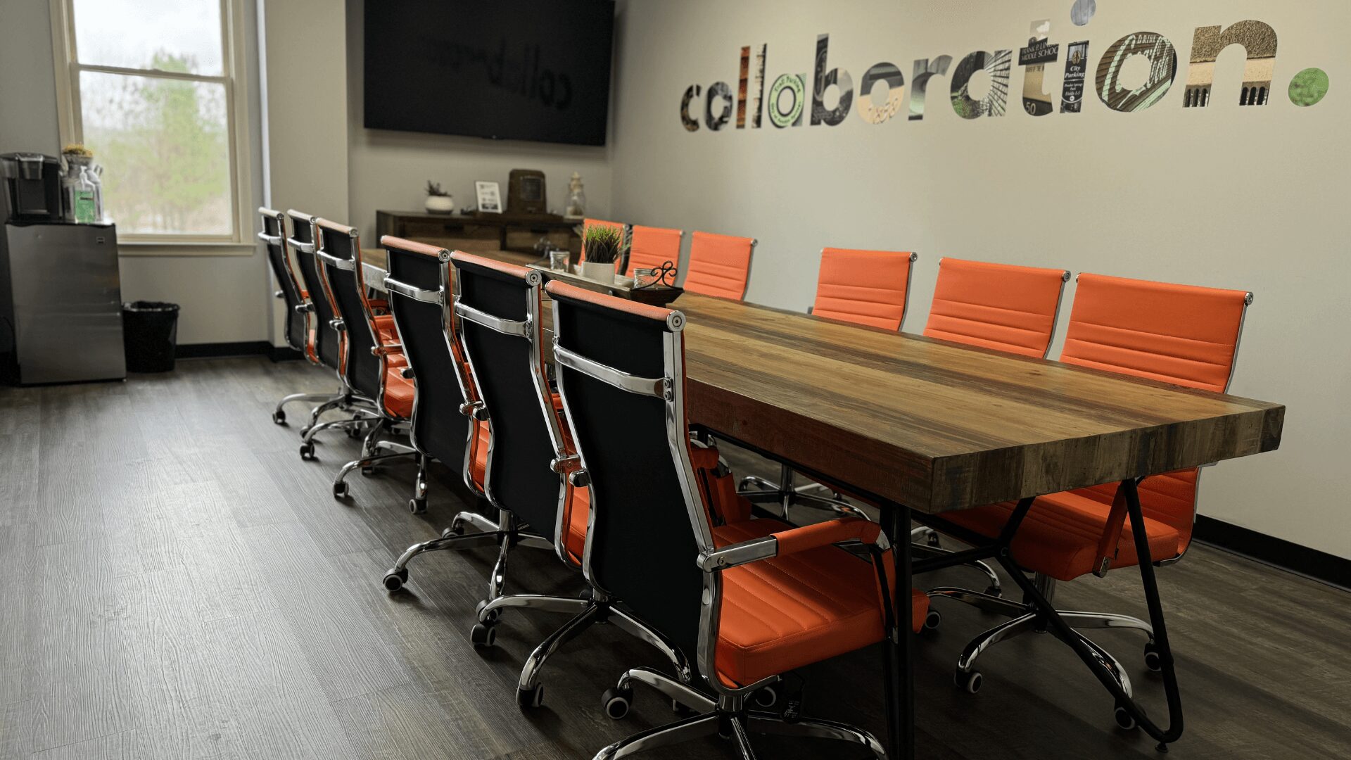 Conference room with a long wooden table, orange chairs, a wall with the word 'collaboration' and a window.