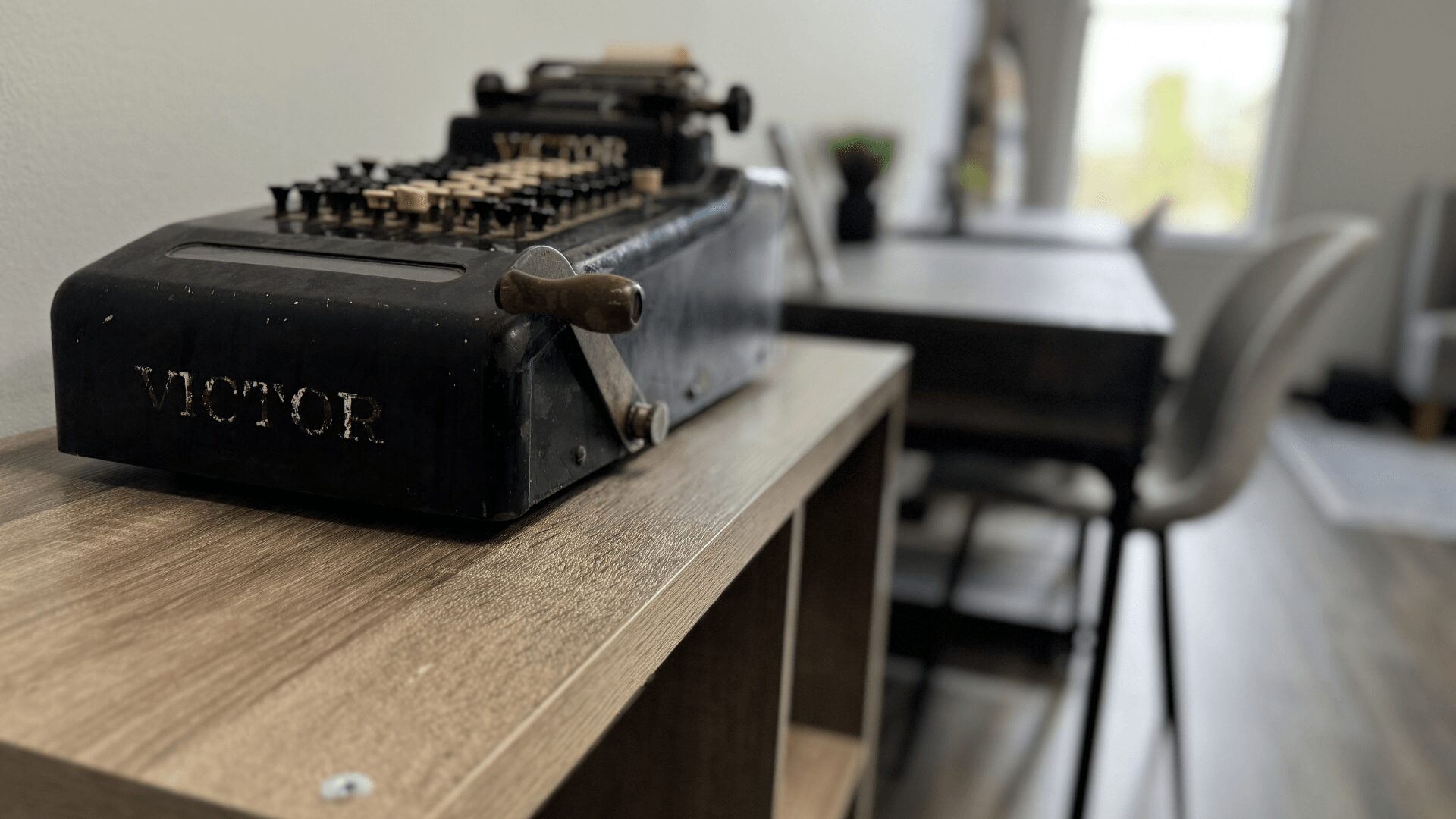 Typewriter on a wooden table in a room with chairs and a window in the background.