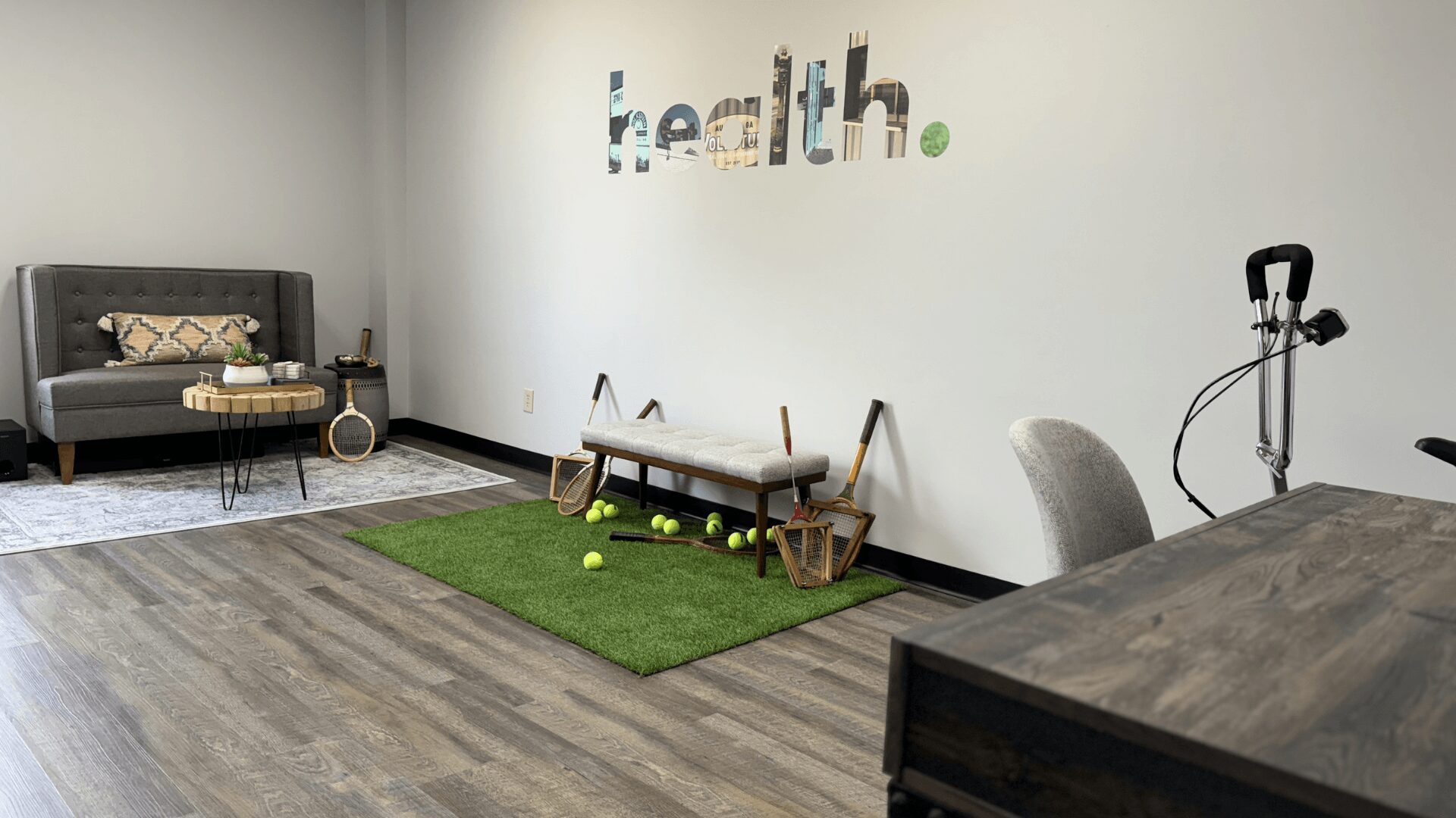 Room with a black sofa, wall art spelling 'health', exercise equipment, and a small green mat with tennis balls.