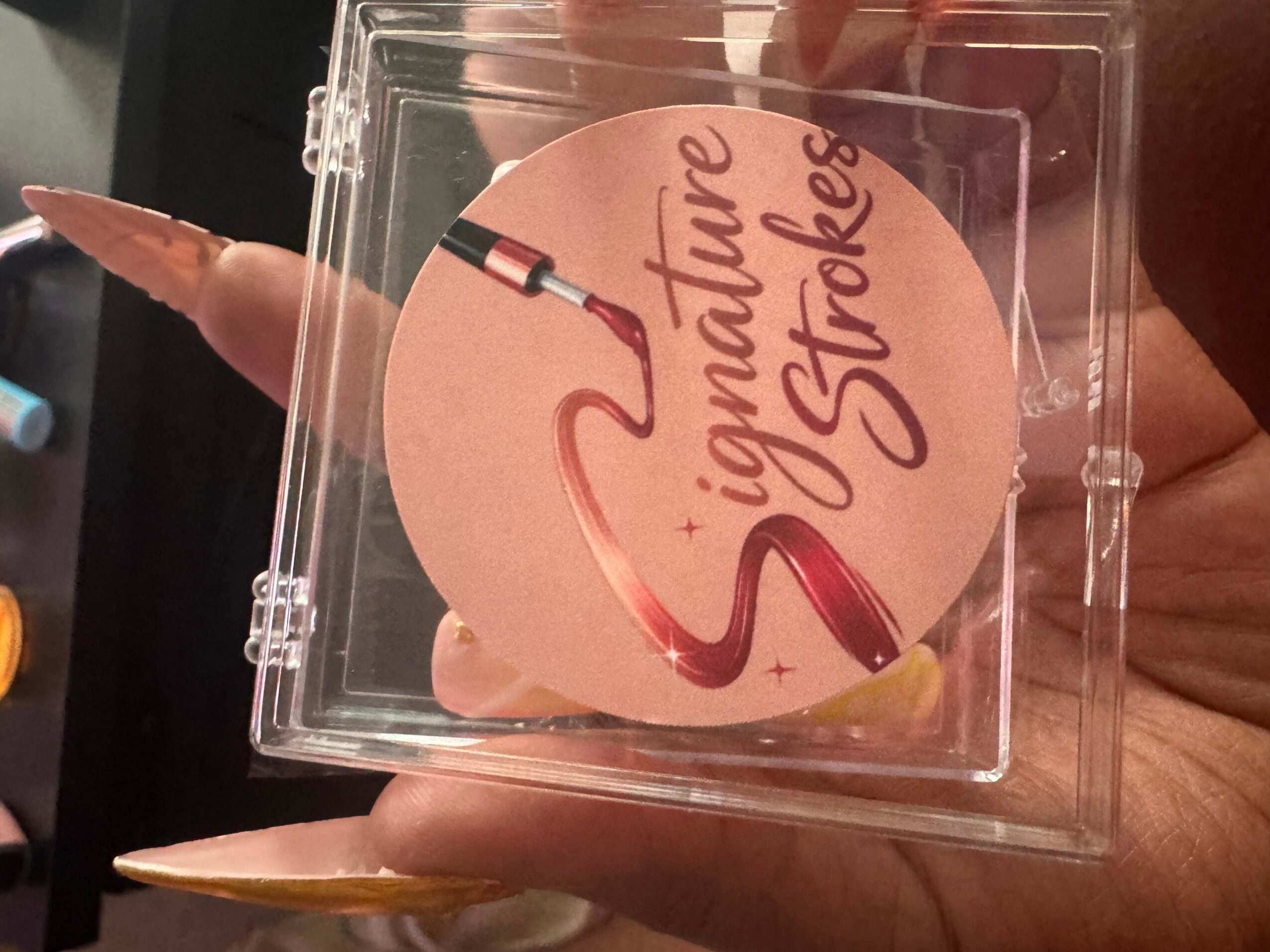 A pink circular card with the words 'ignature Strokes' and a paintbrush illustration inside a clear plastic case.