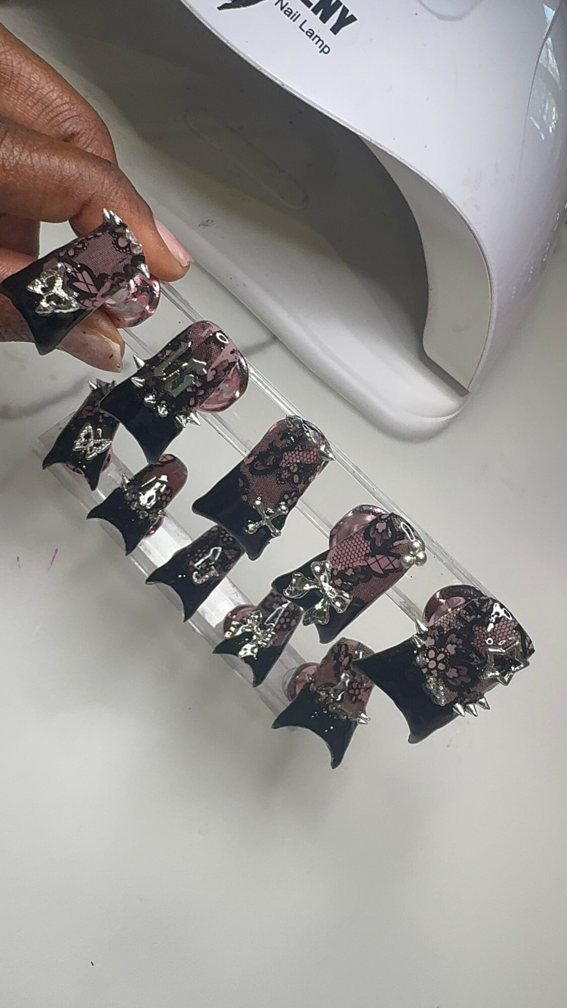 Person's hand with rings holding decorated false nails, with nail dryer in background.