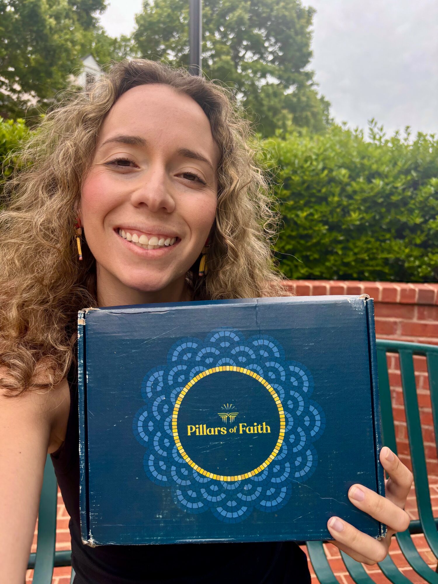 Smiling woman with curly hair holding a blue box labeled 'Pillars of Faith' outdoors.