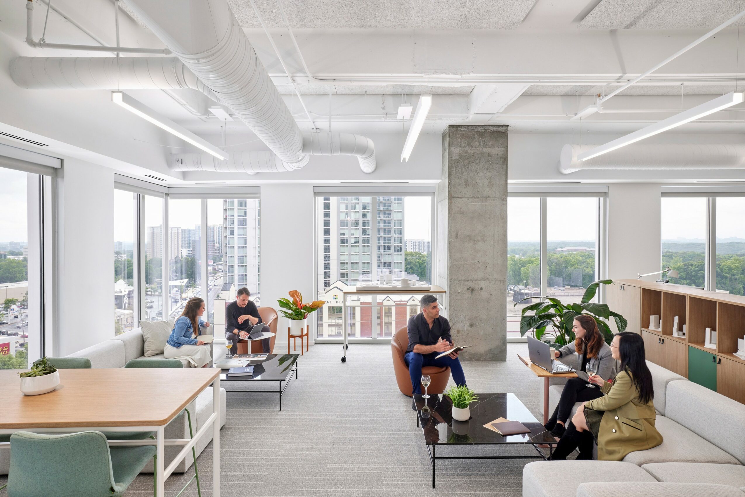 People sitting and working in a bright, modern office with large windows and city views, plants, and contemporary furniture.