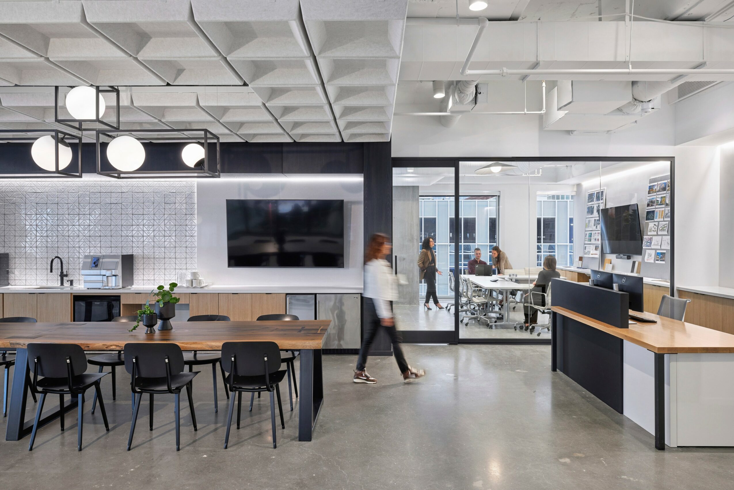 Modern office space with a large TV, conference table, chairs, and a glass wall showing outdoor city view.