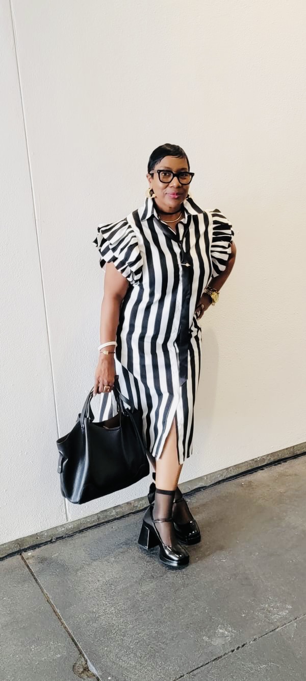 Woman in striped dress with puffed sleeves, glasses, holding a black bag, standing against a plain white wall.