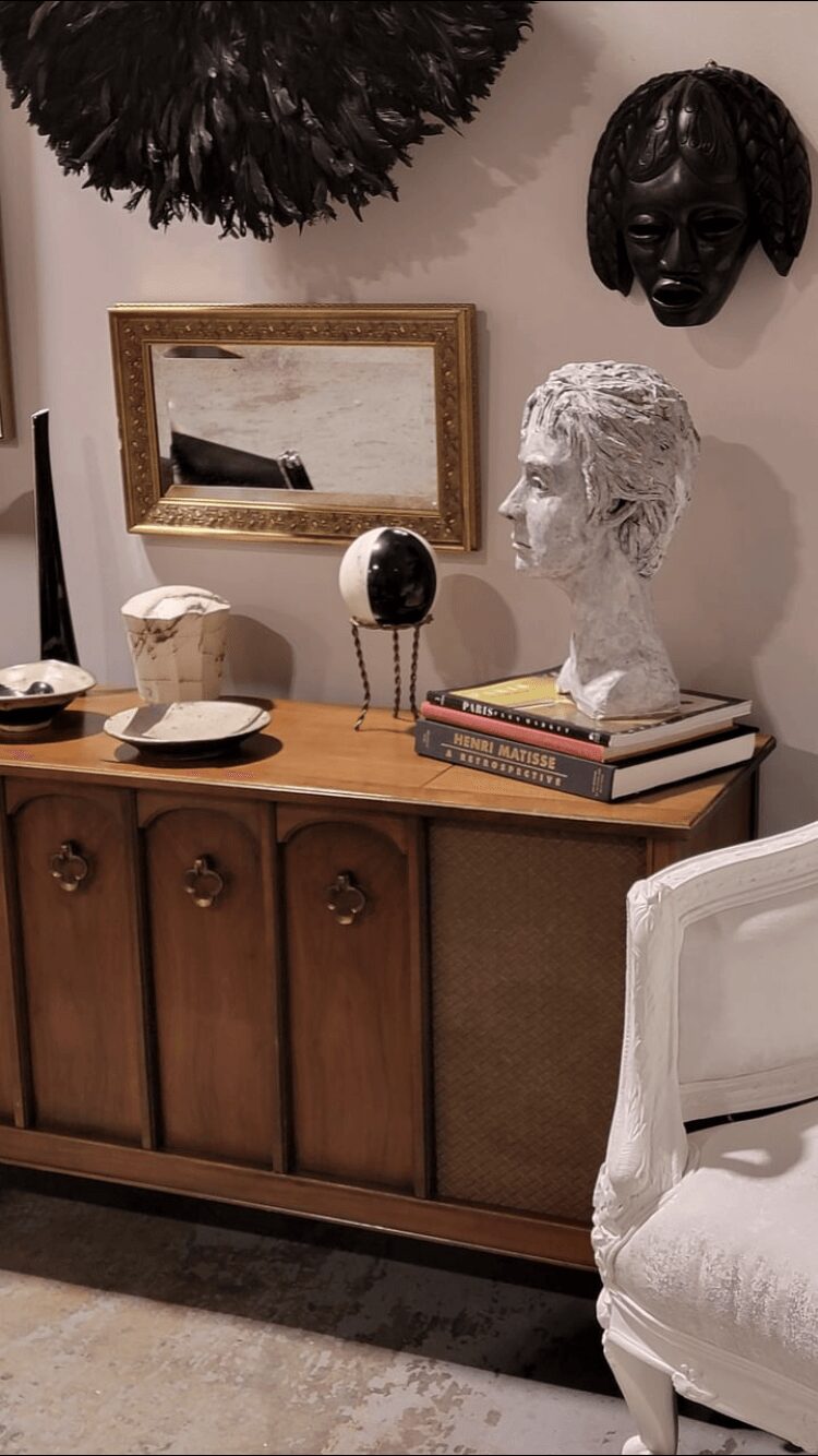 Decorative items on a wooden sideboard, including a bust, mirror, books, and sculptures, with wall art above.
