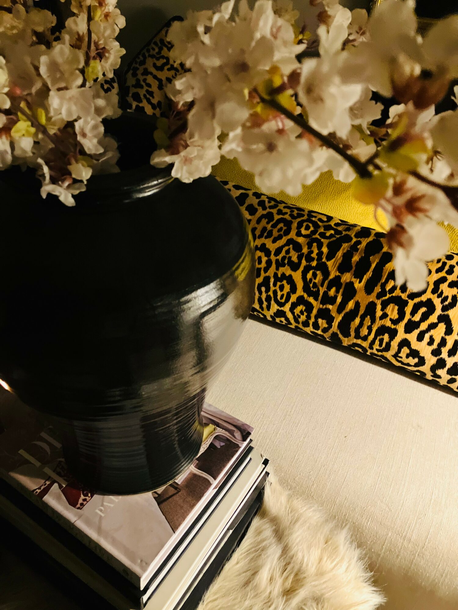 Black vase with white flowers, leopard print object, and a cream-colored surface with a furry object nearby.