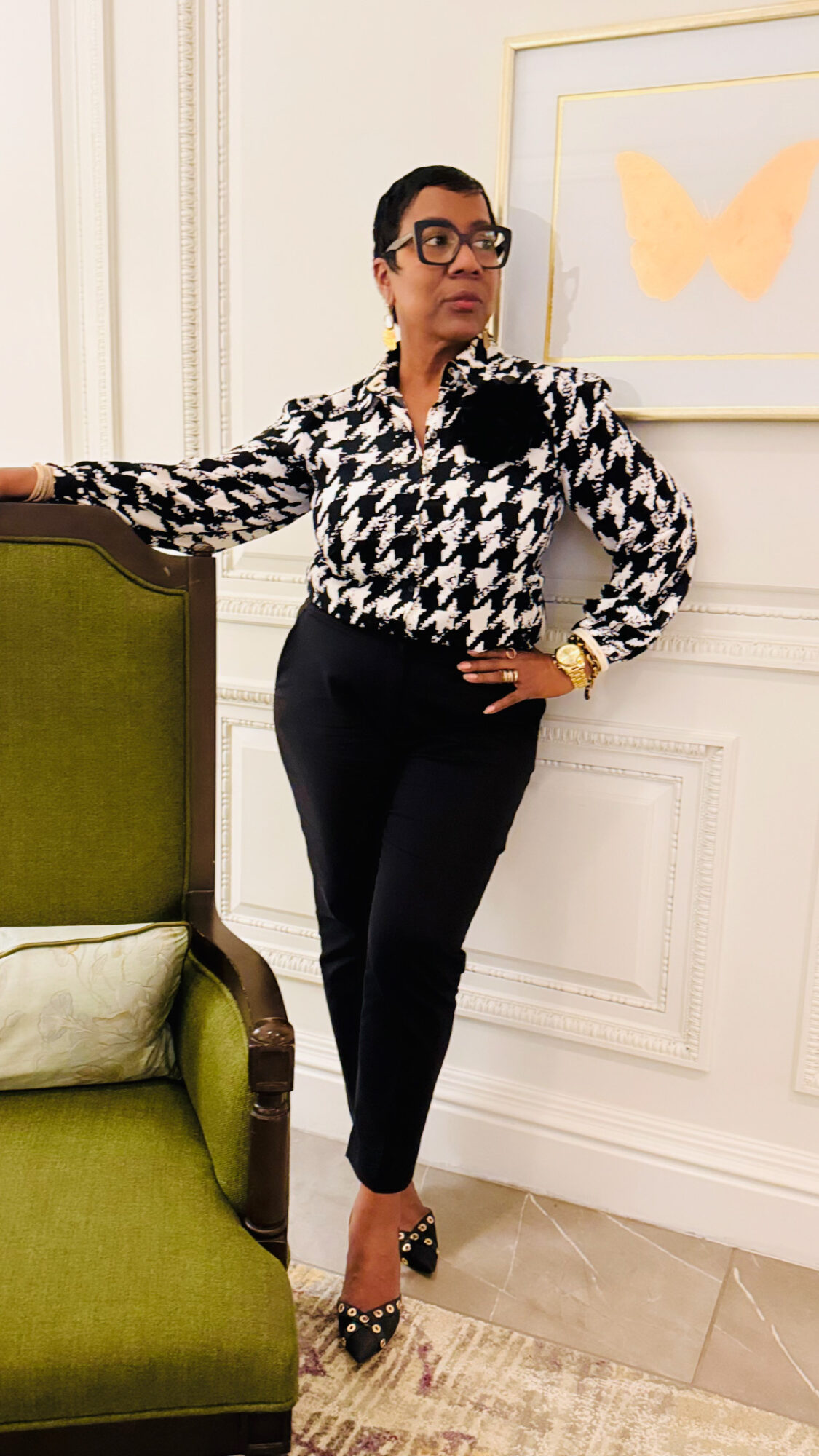 Woman with short hair and glasses stands with one hand on a green chair, wearing a black and white patterned blouse and black pants.