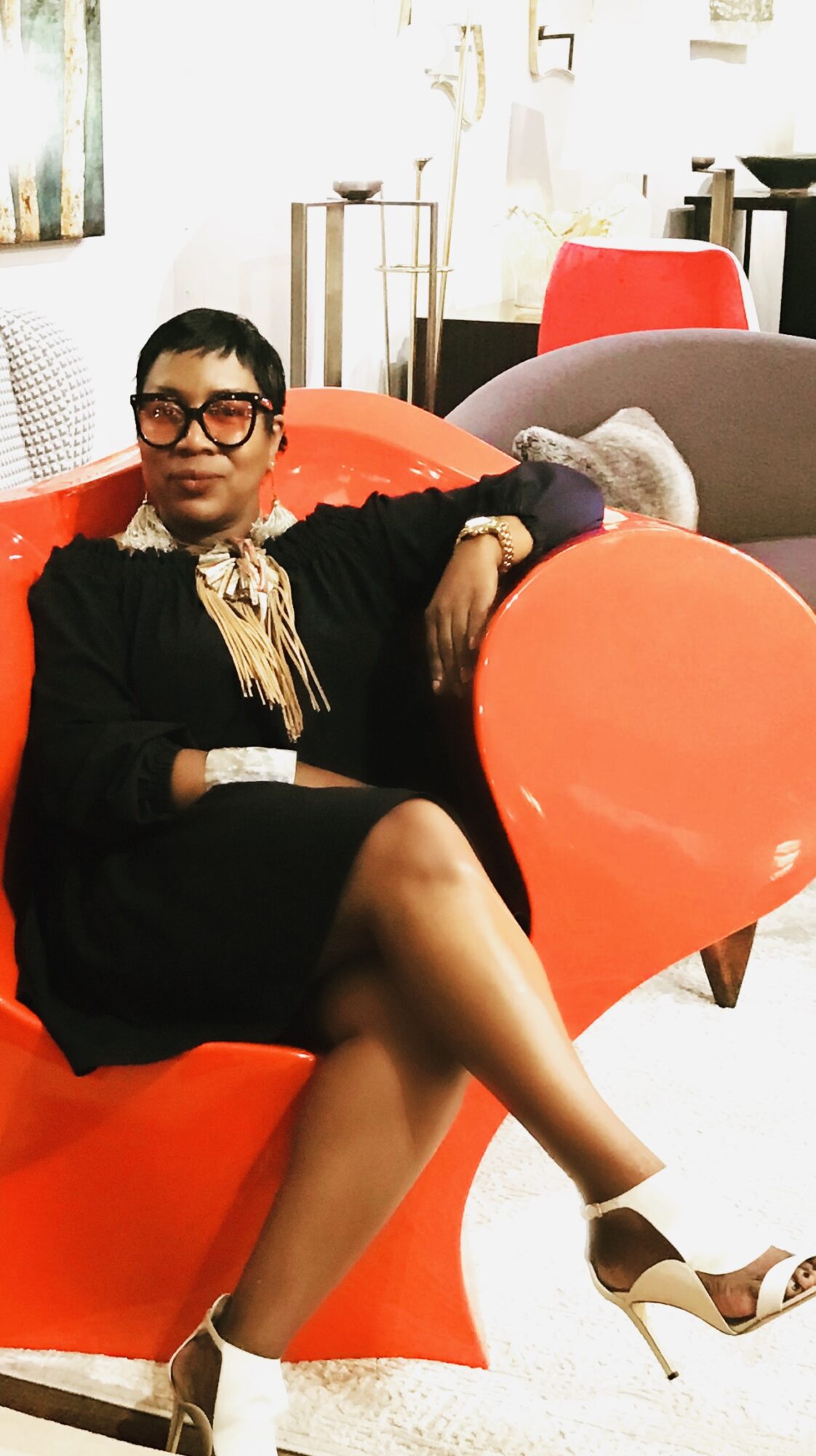 Woman sitting on a red sofa, wearing glasses, a black dress, and high heels, in a bright room.