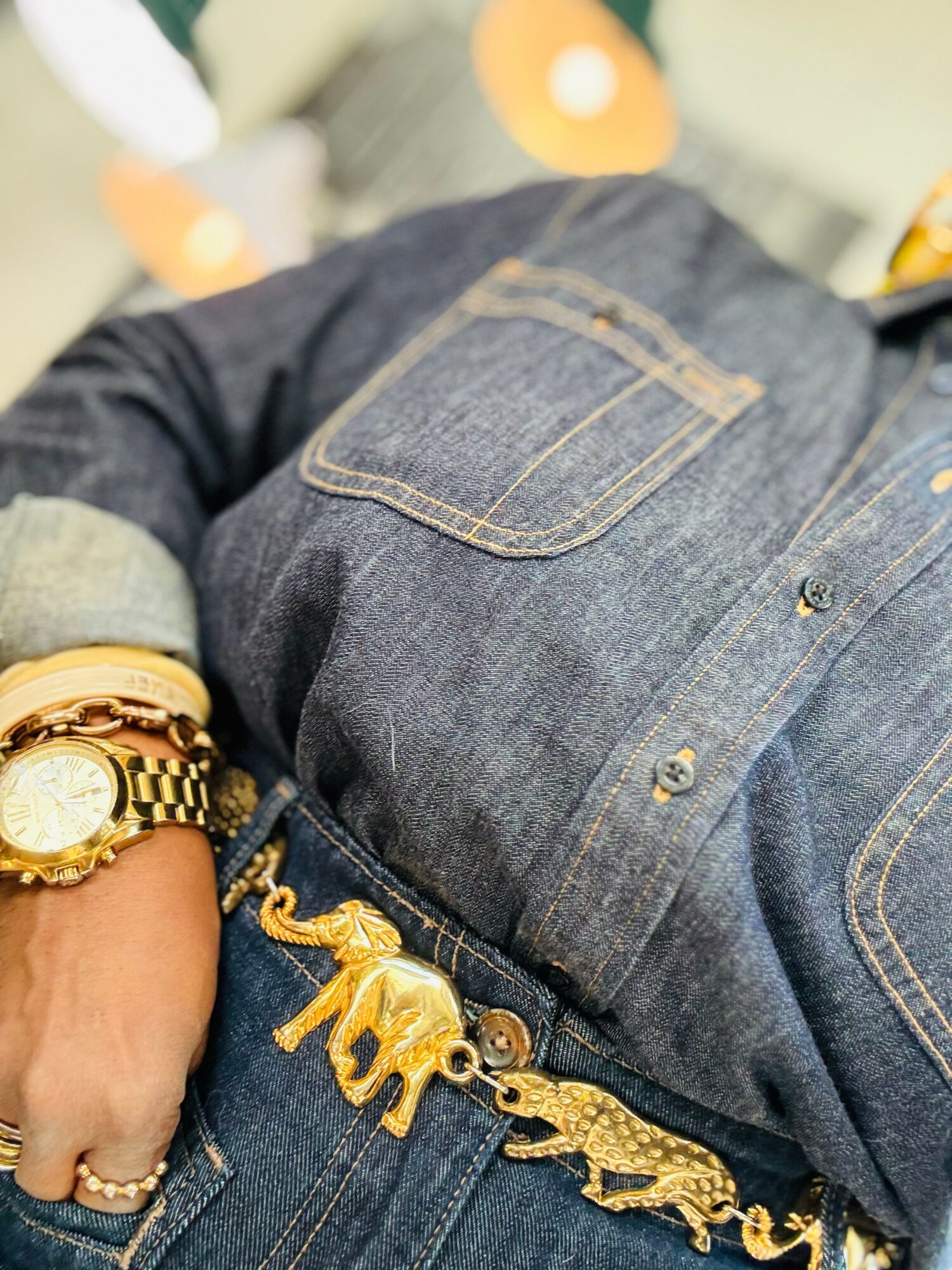 Close-up of a person wearing a denim shirt with a pocket, gold jewelry, and a belt with an elephant and crocodile charm.