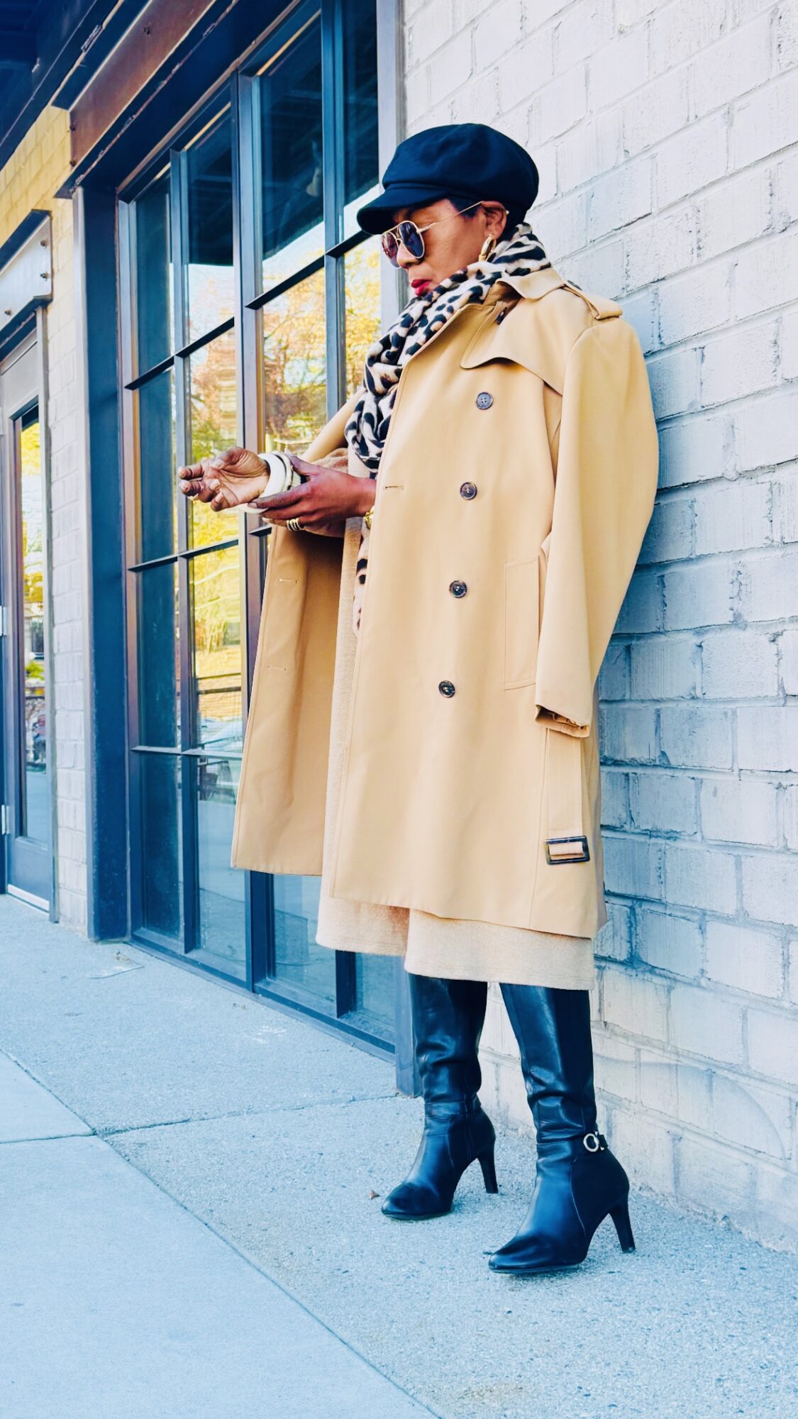 Person in beige trench coat, black hat, and high heels stands outside, looking at their phone near a blue storefront.