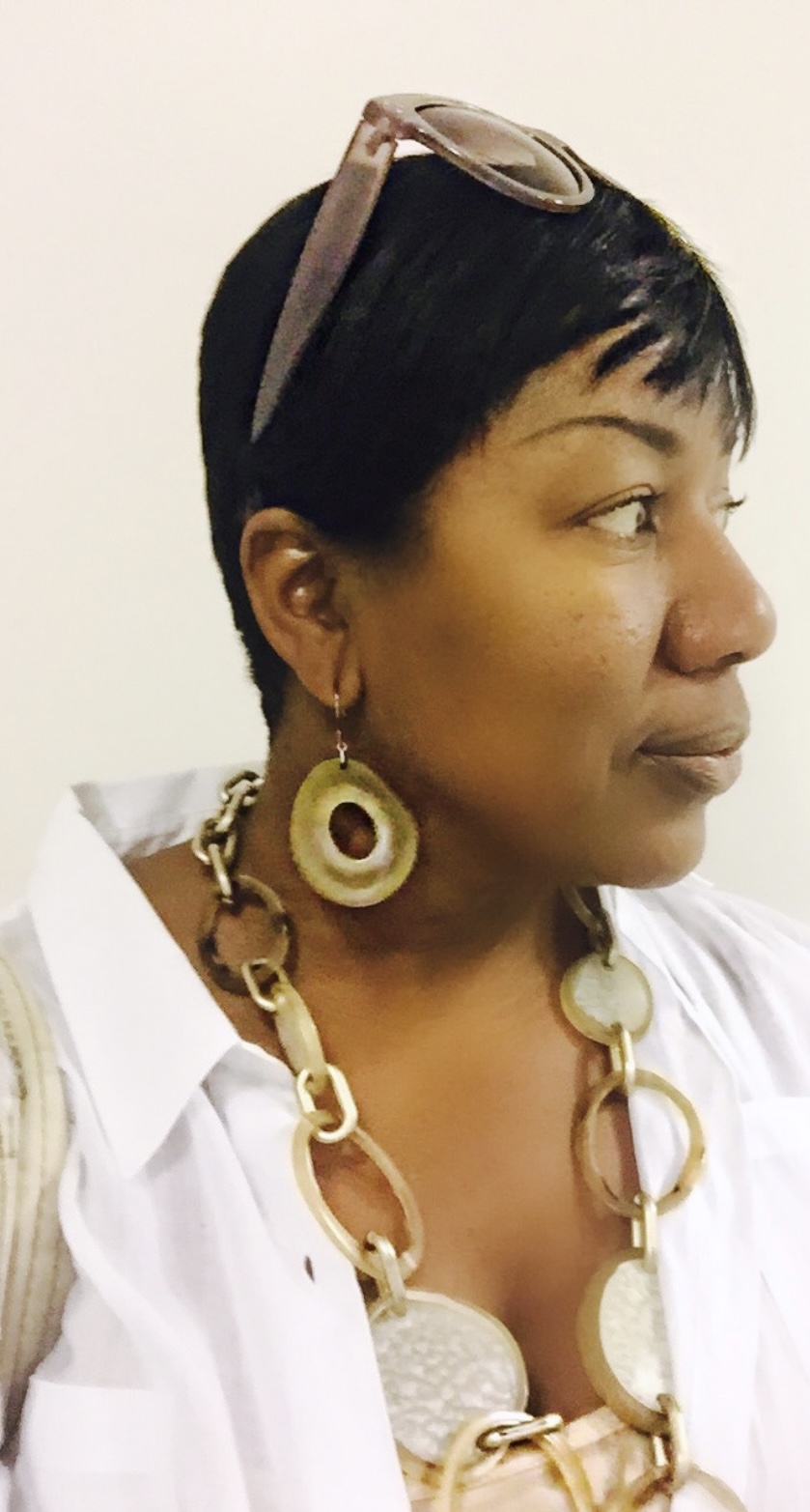 Side profile of a woman with short black hair, wearing large earrings, a chunky necklace, and sunglasses on her head.