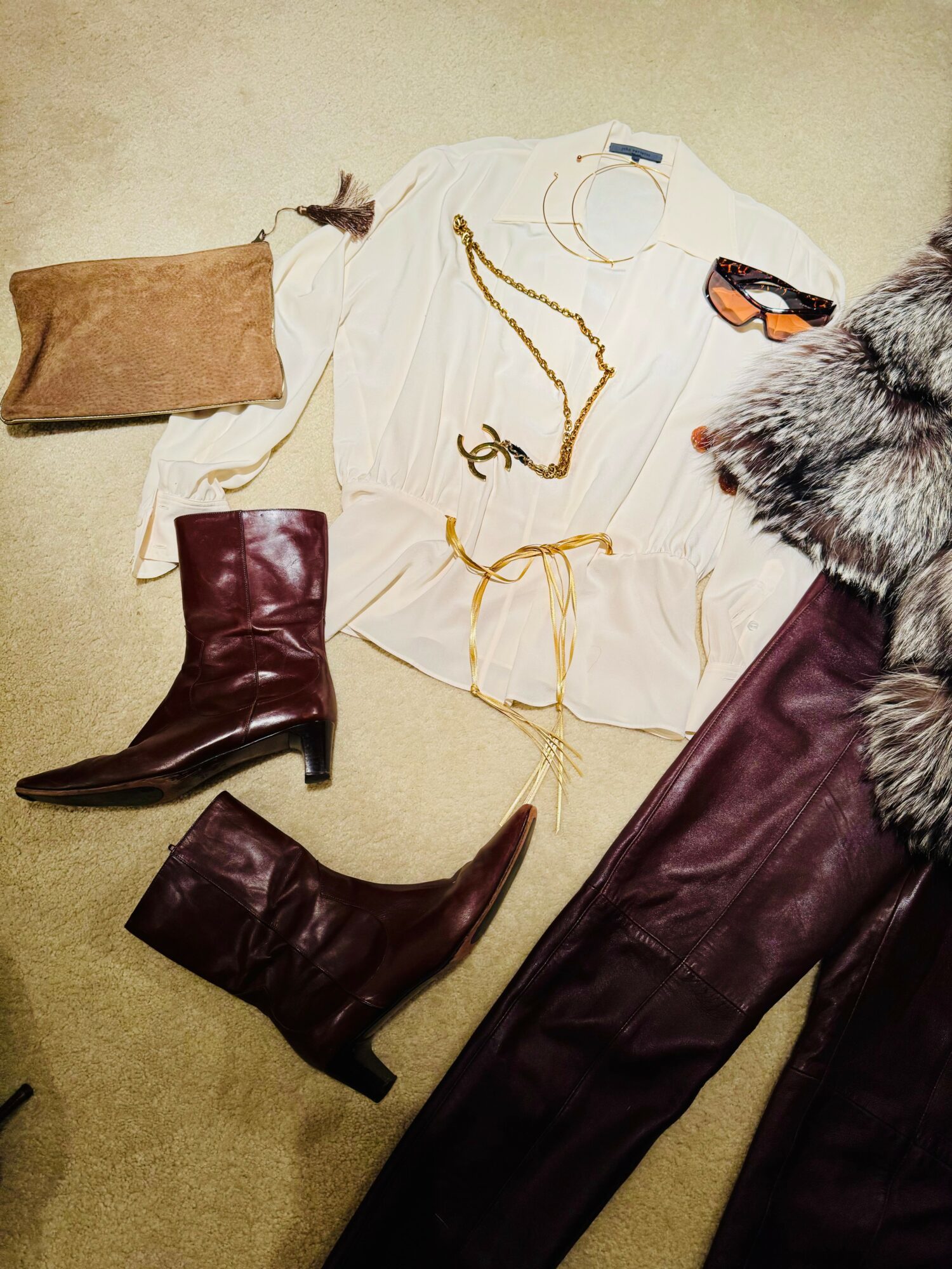 Flat lay of clothing and accessories including a white shirt with gold jewelry, brown boots, a beige clutch, sunglasses, and a fur coat.