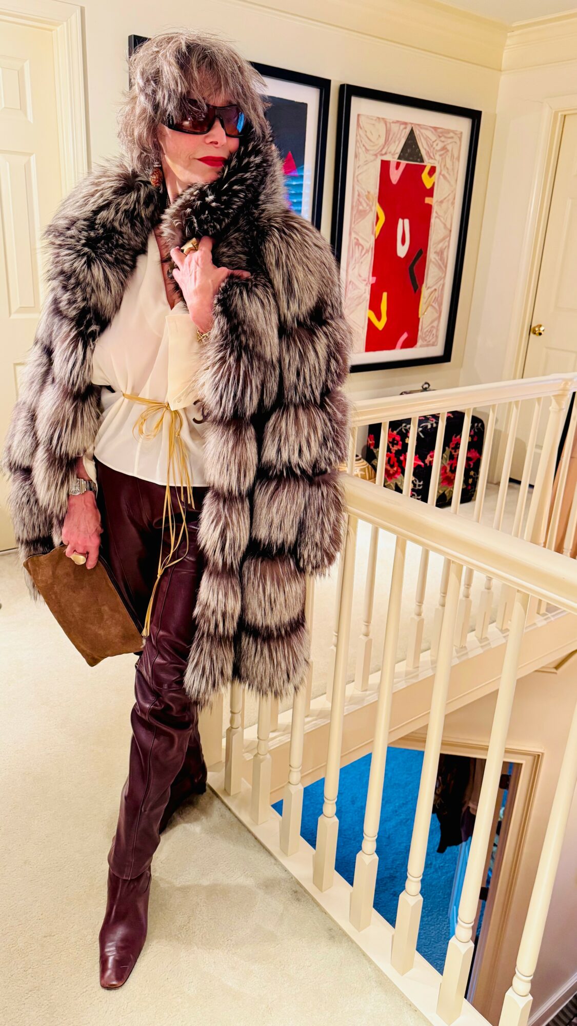 Person in fur coat and sunglasses standing indoors near a staircase, holding a bag, with framed artwork on the wall.