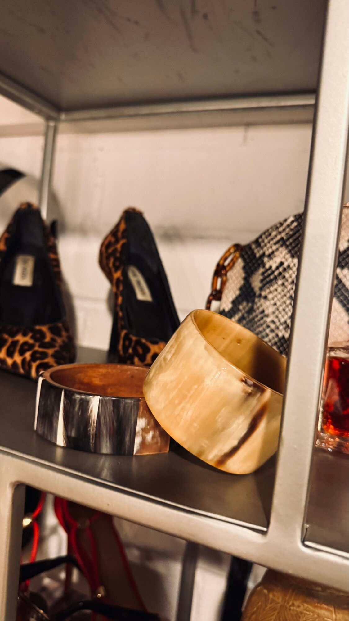 Two bracelets, one metallic and one wooden, placed on a shelf with shoes and a bag in the background.