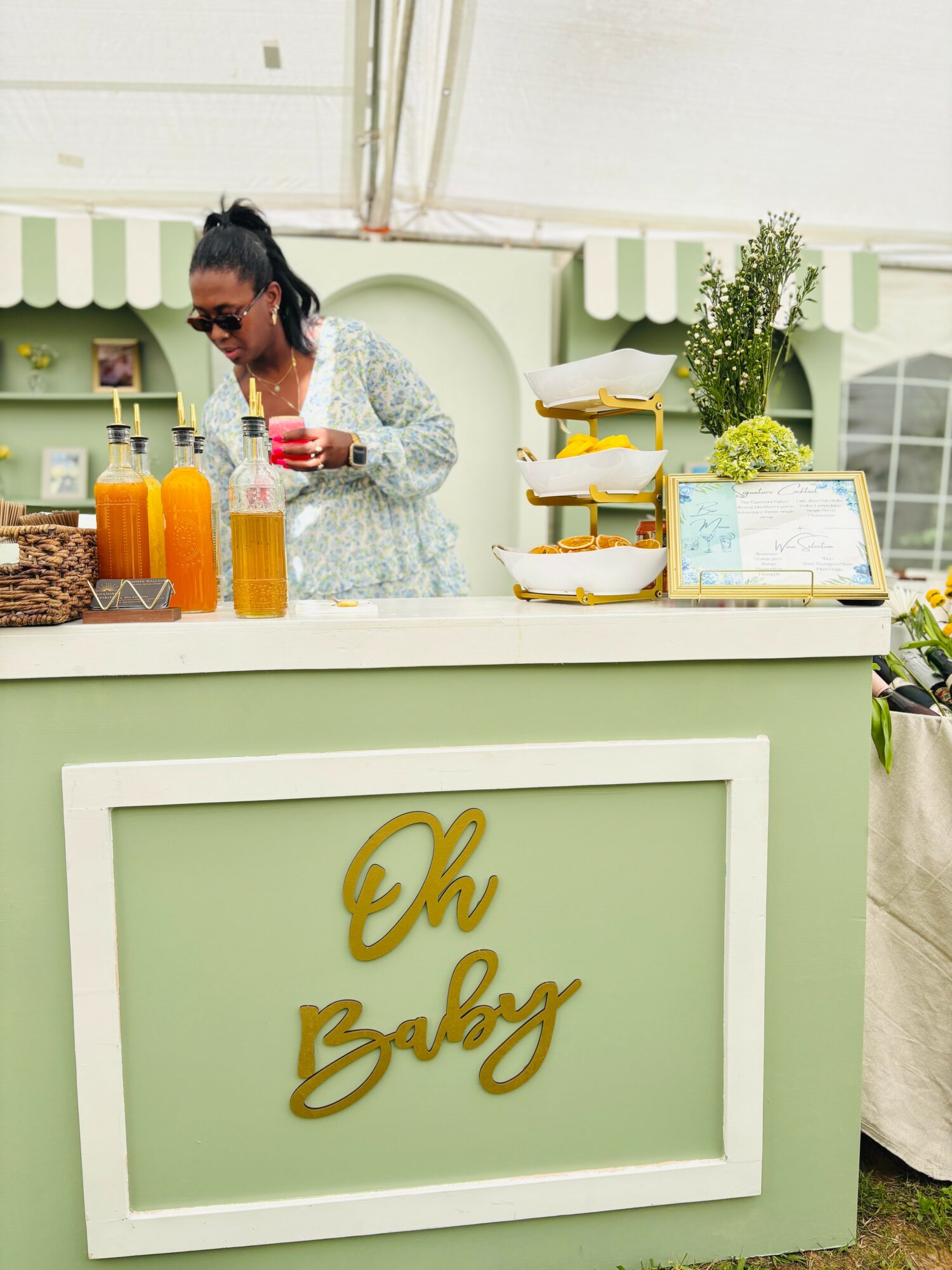 Woman in sunglasses at a green and white booth with drinks and snacks, sign reads 'Oh Baby'.