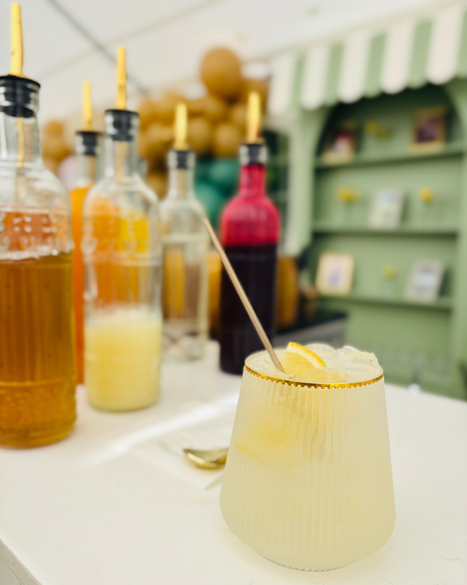A yellow drink with a straw and lemon slice in a glass, with bottles and a green shelf in background.