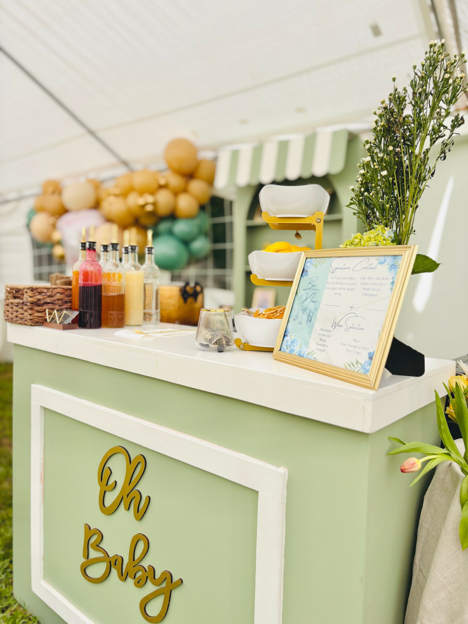 Counter with a green and white striped awning, decorated with flowers, bottles, and a framed menu, in a bright setting.