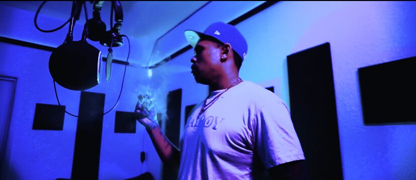 Person in a room with blue lighting, holding a vape device, wearing a cap and t-shirt, surrounded by soundproof panels.