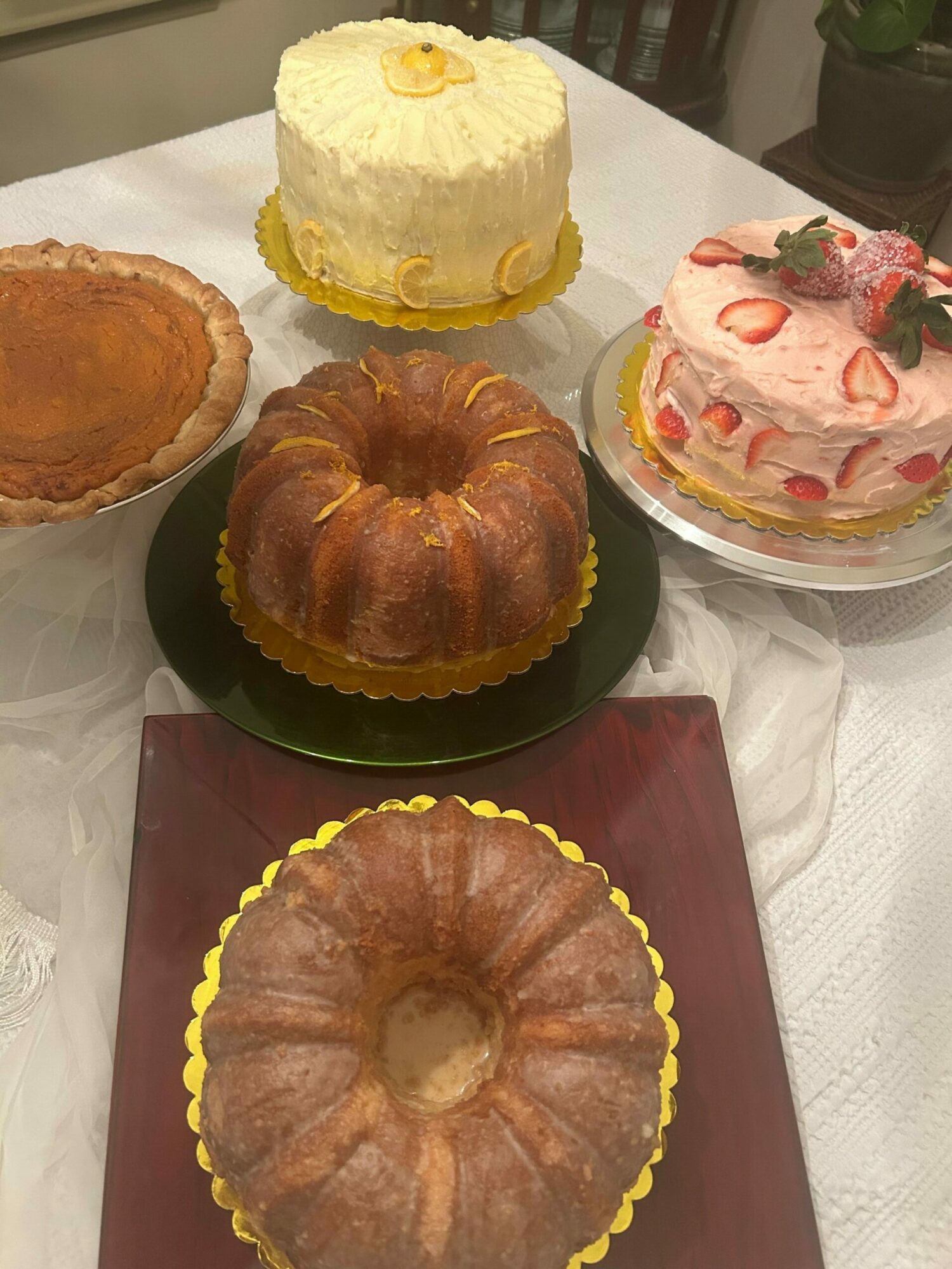Four desserts on a table: a cake with pink and white frosting, a yellow cake, a bundt cake, and a small round cake, all on different plates or stands.