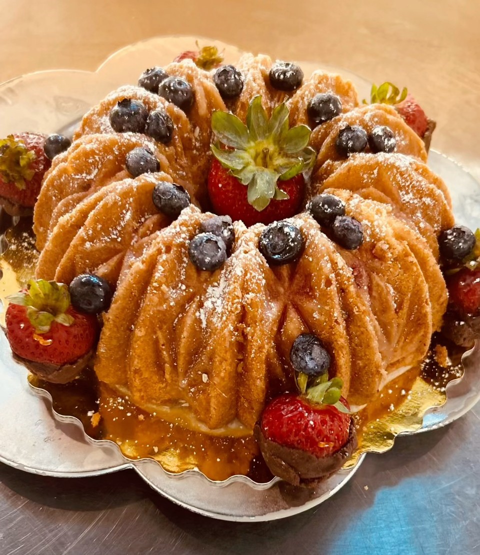 Bundt cake decorated with strawberries, blueberries, and powdered sugar, on a silver platter, with caramel sauce drizzled around.