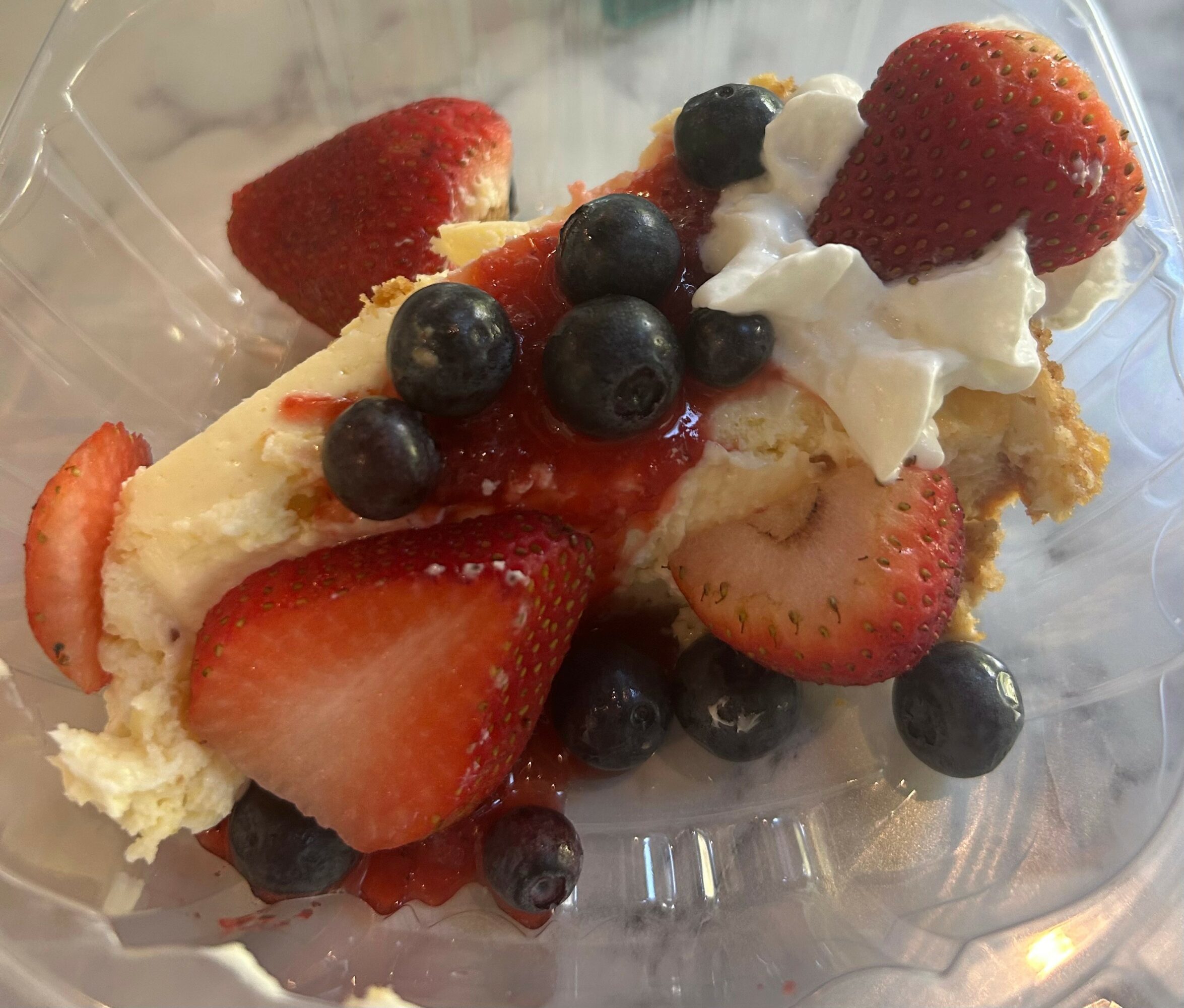 Slice of cake topped with strawberries, blueberries, and whipped cream, in a clear container.