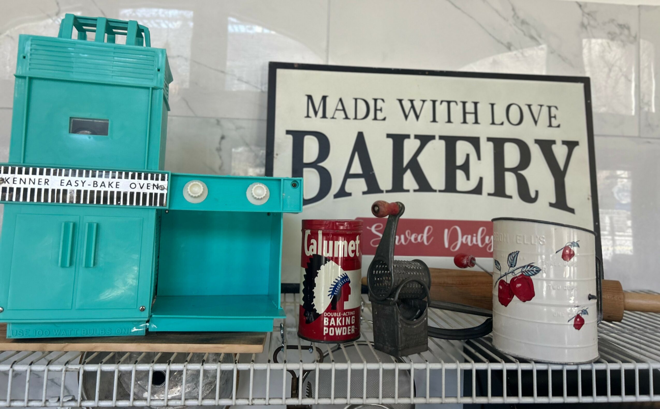 Bakery display with teal oven, sign, red syrup bottle, black kitchen tool, and white container with cherries, on a wire shelf.