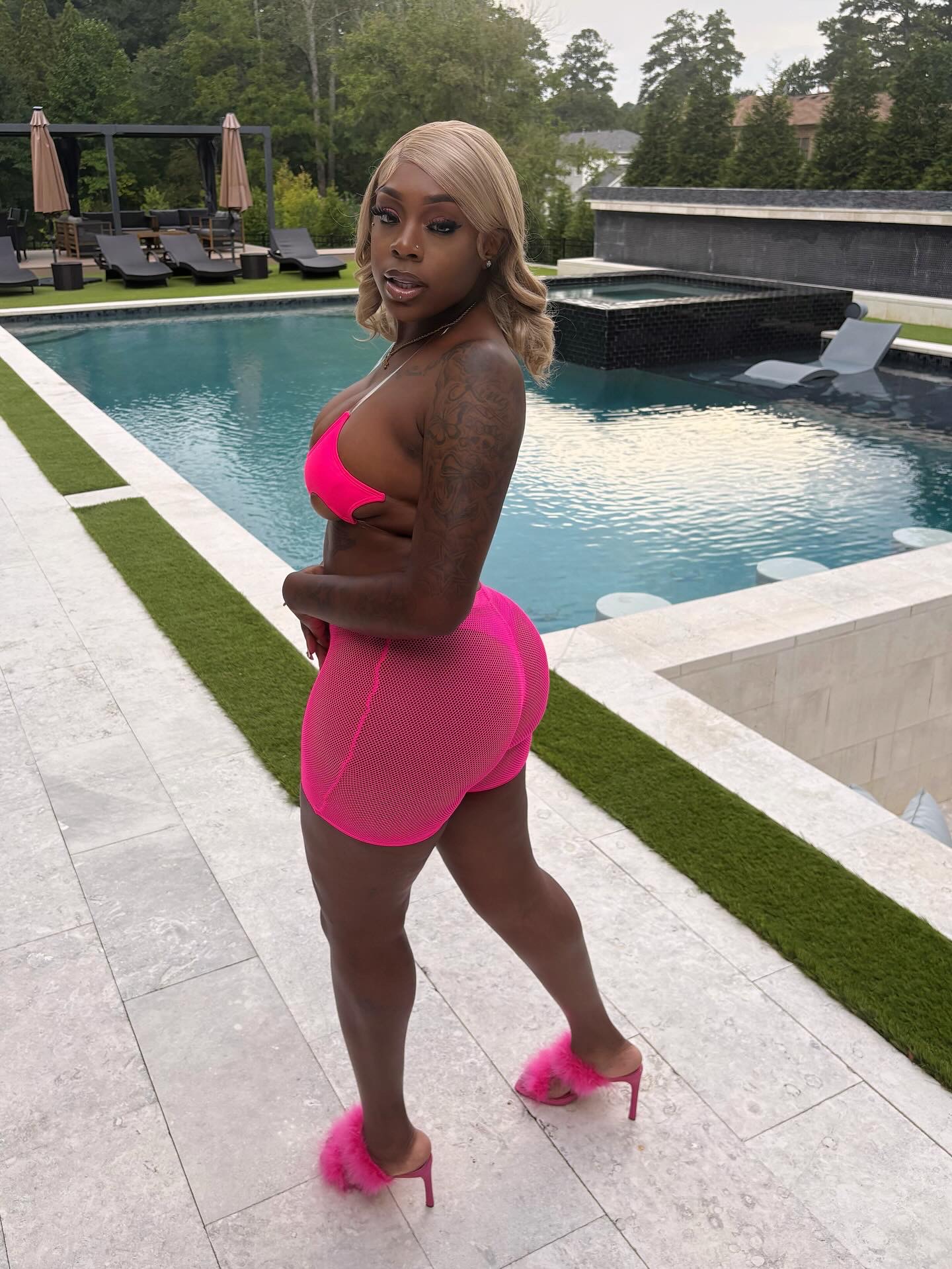 Woman in pink dress and high heels standing by poolside, with pool and outdoor furniture in background.